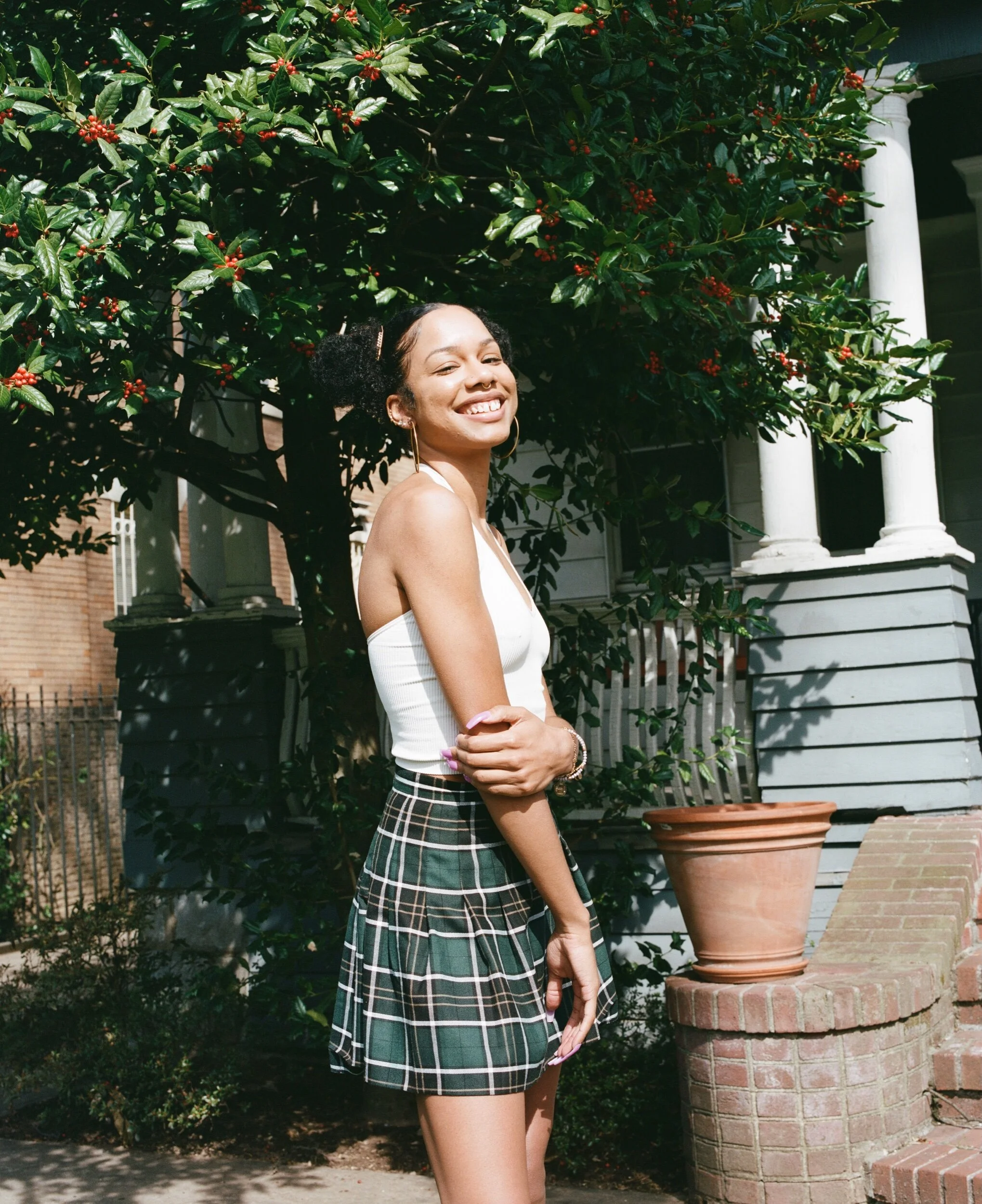 Smiling woman with dark curly hair styled in puffs, wearing a white sleeveless top and plaid skirt, standing outdoors with a bush with red berries and residential houses in the background.