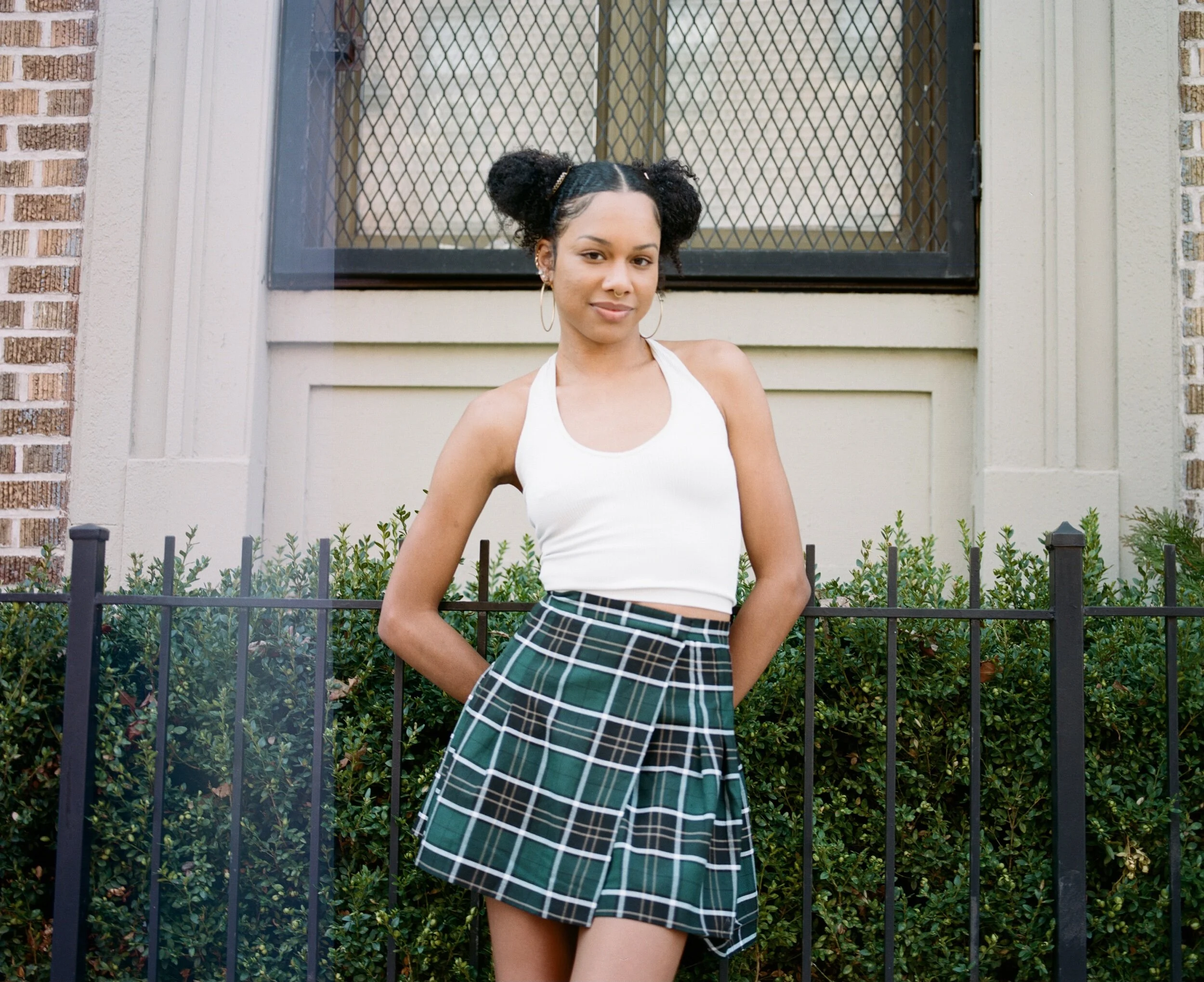 A young woman standing outdoors in front of a black metal fence, wearing a white sleeveless top, a green plaid skirt, hoop earrings, and with her hair styled in puff buns.