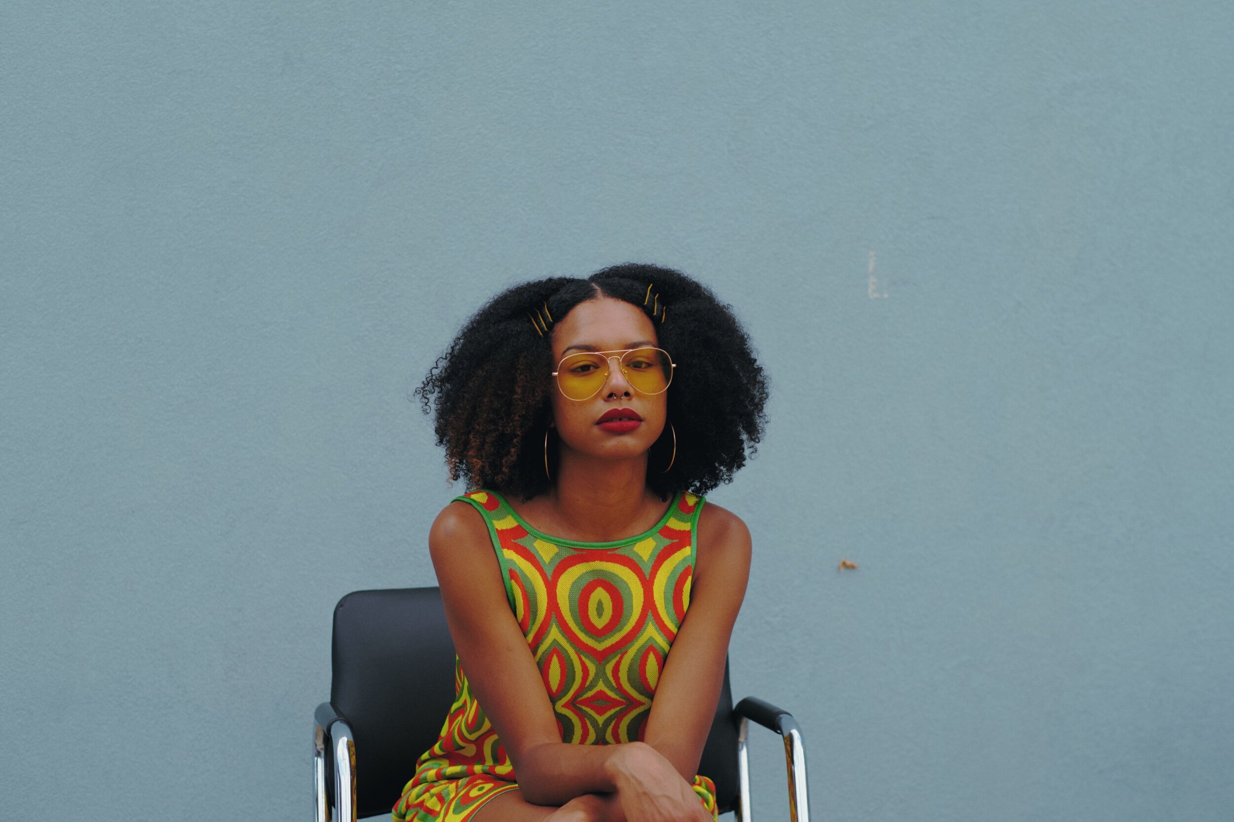 A woman with curly dark hair wearing yellow sunglasses and hoop earrings, sitting on a black chair against a light blue wall, dressed in a sleeveless dress with a colorful swirl pattern.