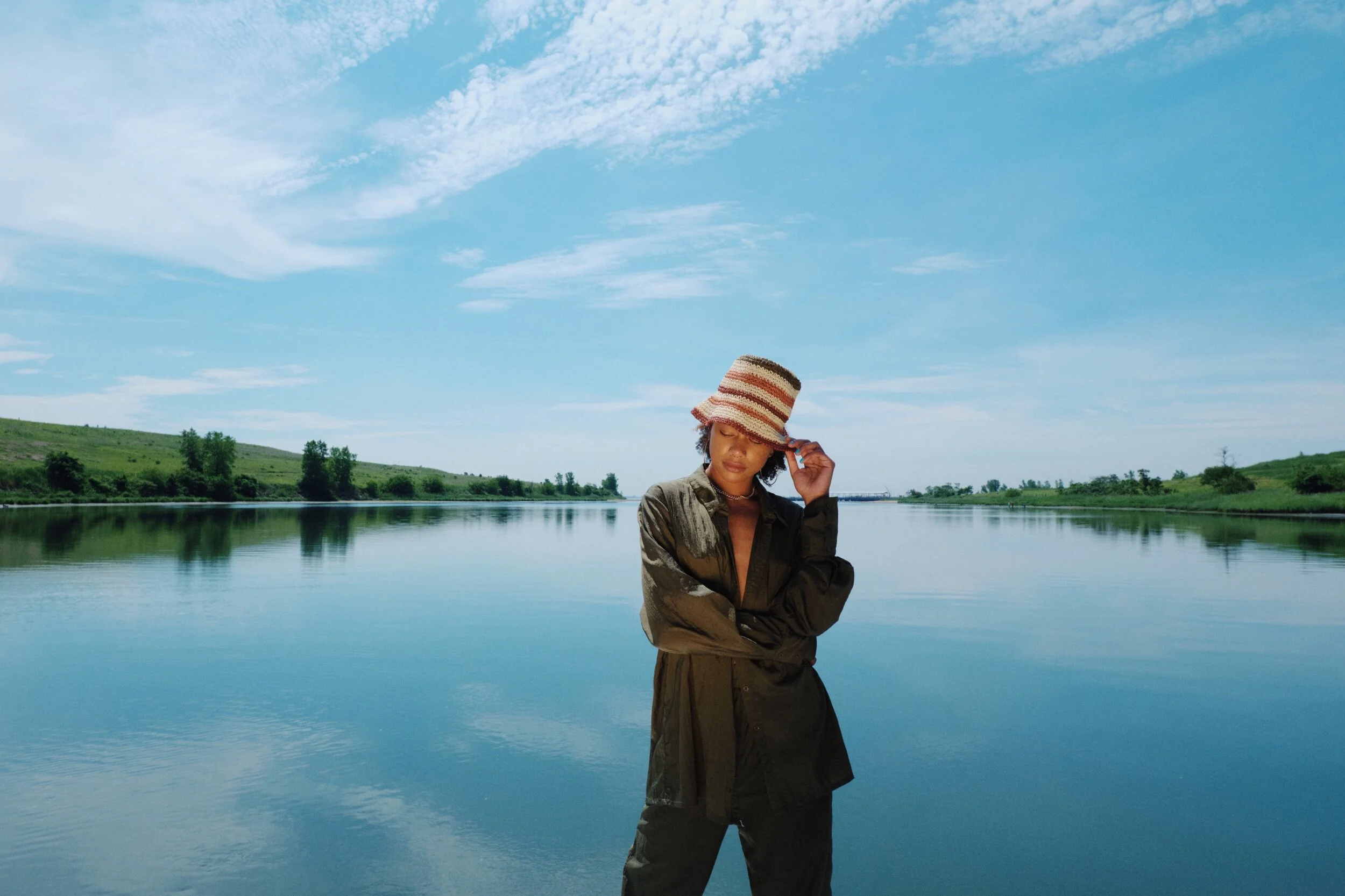 A woman wearing a striped wide-brimmed hat and a dark jacket standing beside a calm river with green hills and blue sky in the background.