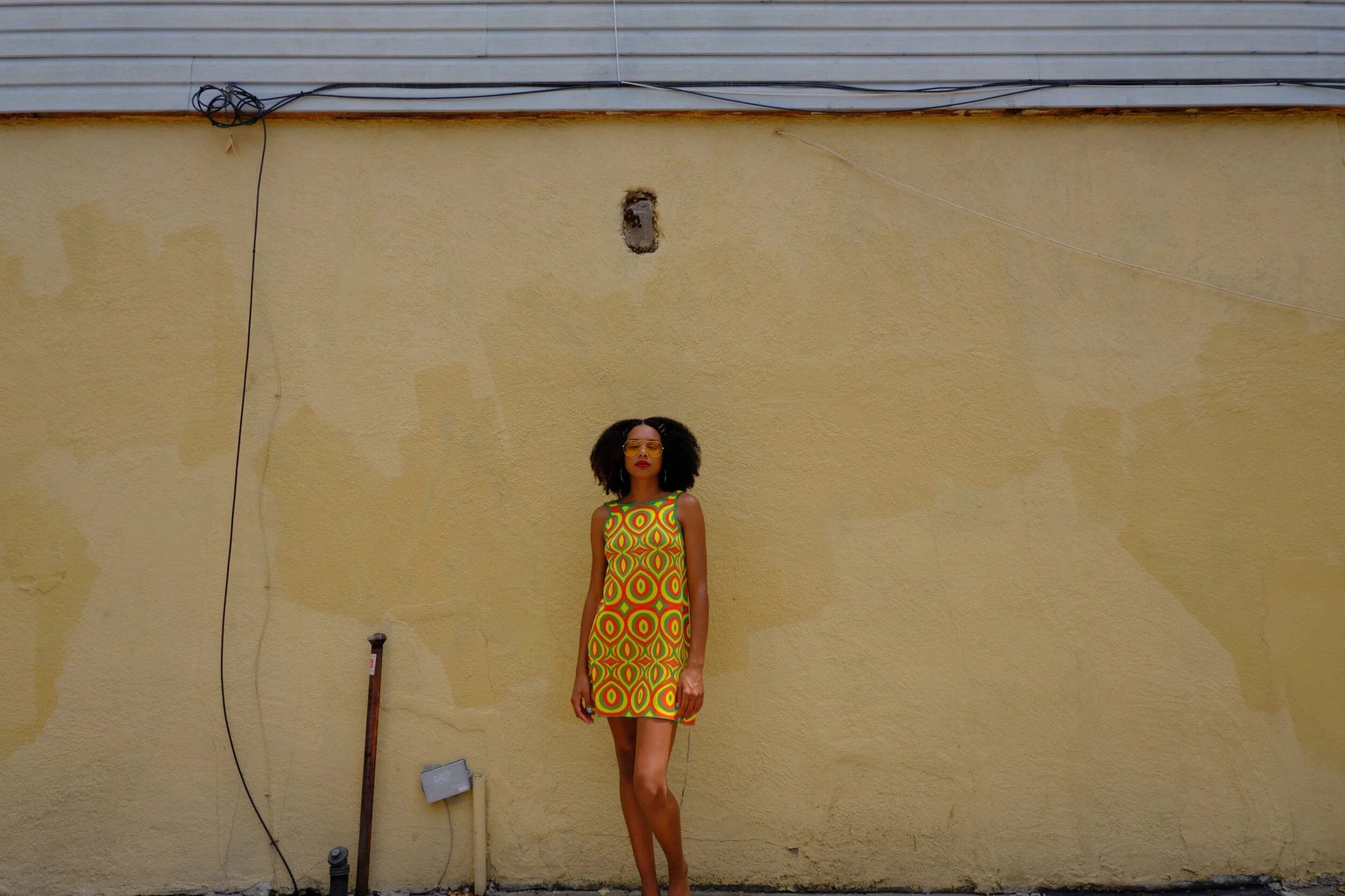 A woman with curly hair wearing yellow glasses and a colorful, geometric patterned dress standing in front of a yellow wall.
