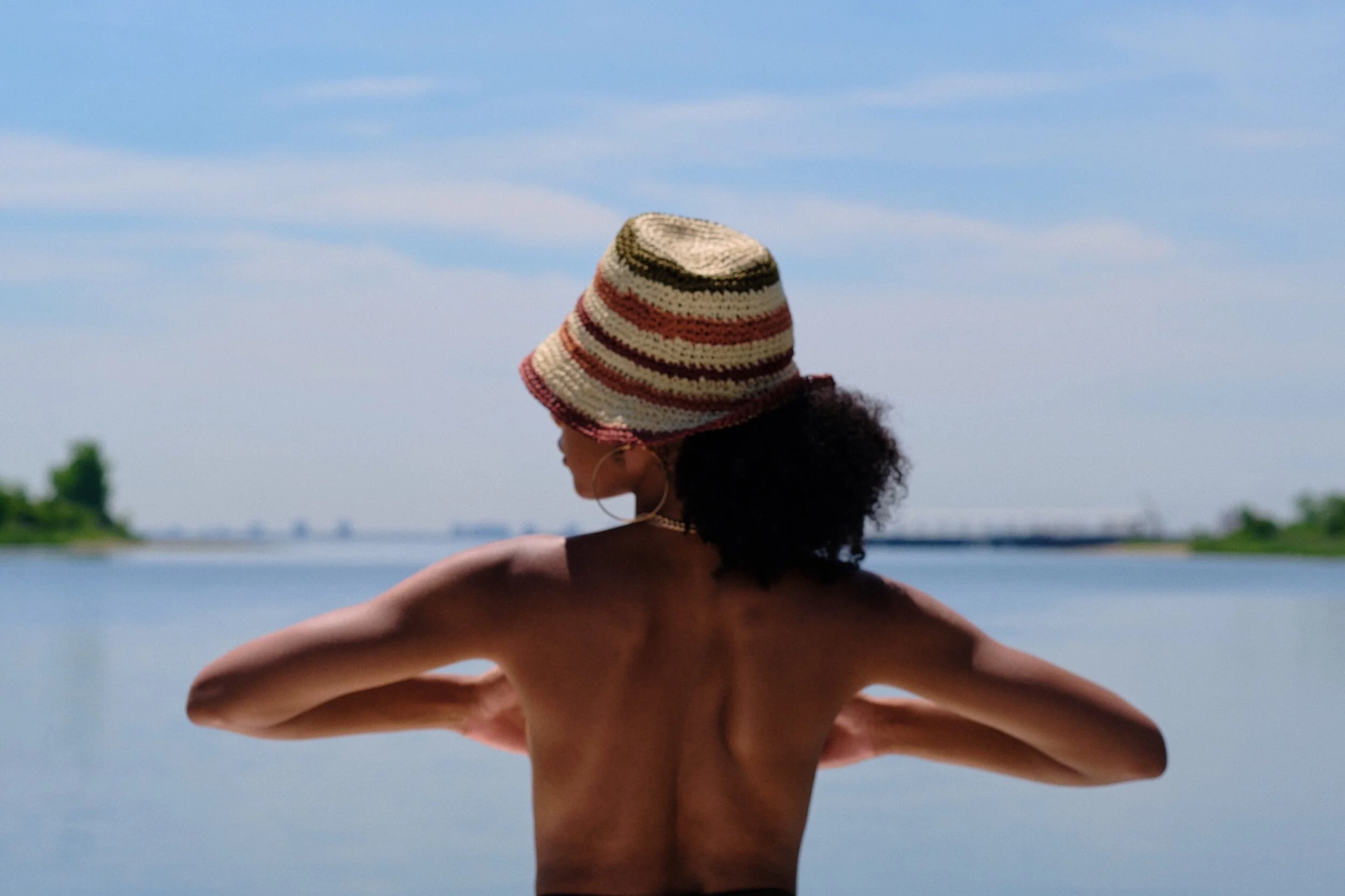 A woman with curly hair wearing a striped crochet hat, facing water and sky, with her arms outstretched.