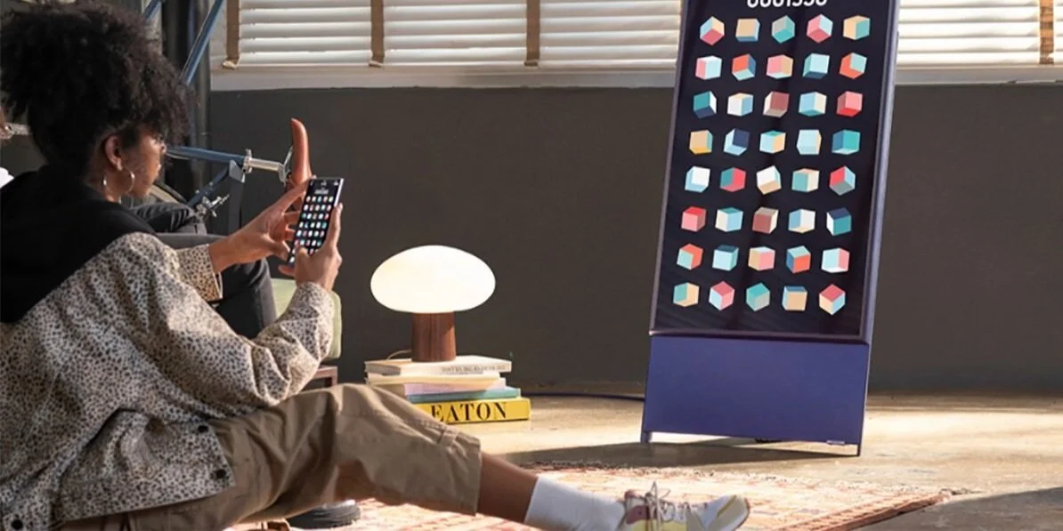 A woman sitting on the floor taking a photo with her phone of a colorful geometric pattern on a display board in a modern, well-lit room with a lamp, books, and a decorative rug.