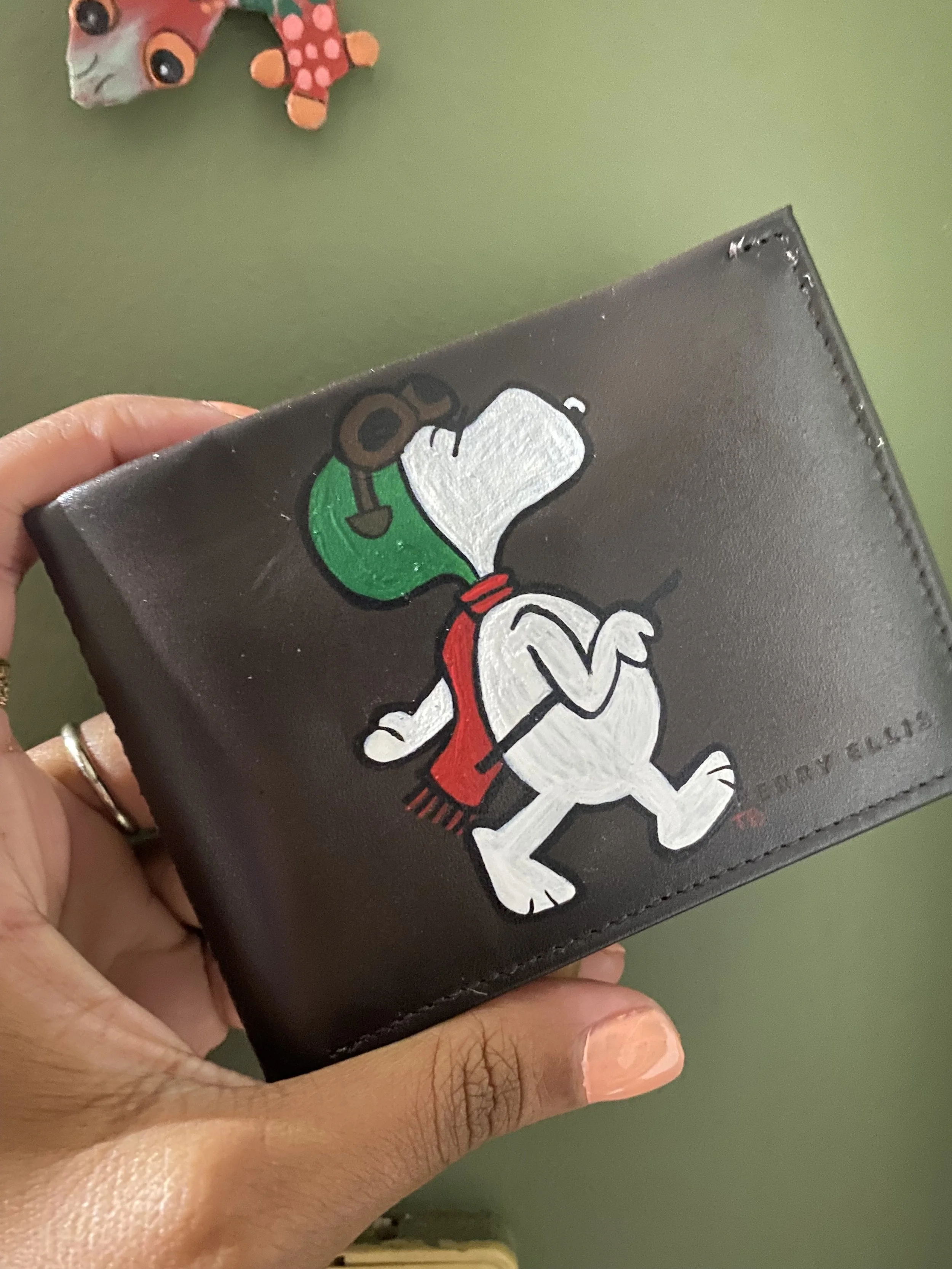 Close-up of a hand holding a black leather wallet with a cartoon sticker of Snoopy wearing a green hat and red scarf on it. In the background, a plush toy character with large eyes, orange hands, and a pink body is visible against a green wall.