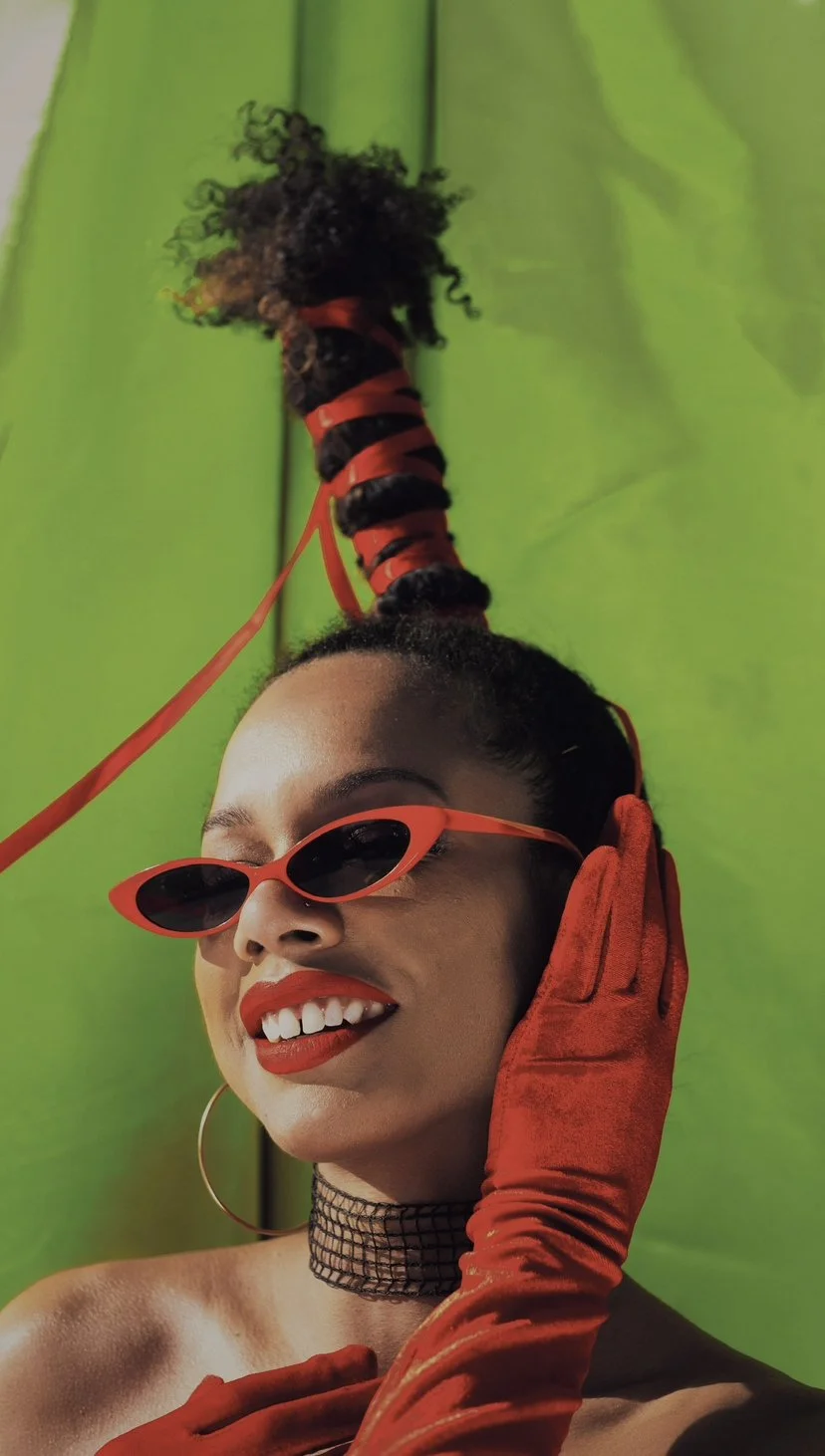 A woman with dark hair styled in a tall curly bun, wearing red sunglasses, red lipstick, a black mesh choker, and red gloves, smiling against a bright green background.