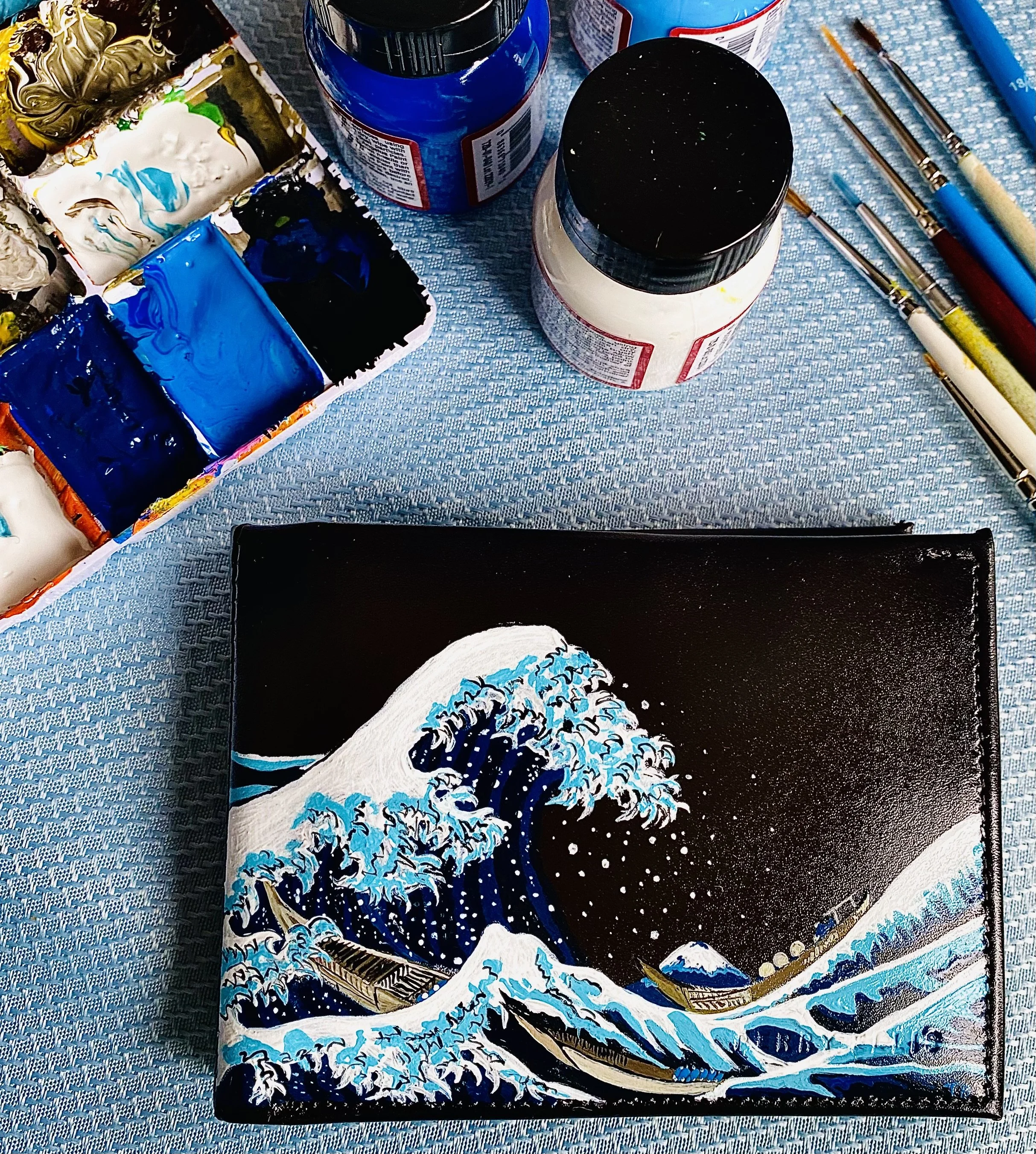 A scene inspired by the famous Japanese artwork The Great Wave off Kanagawa, featuring stylized blue and white ocean waves with foam and spray, set against a dark background. In the foreground, there appears to be a rectangular object resembling a fr