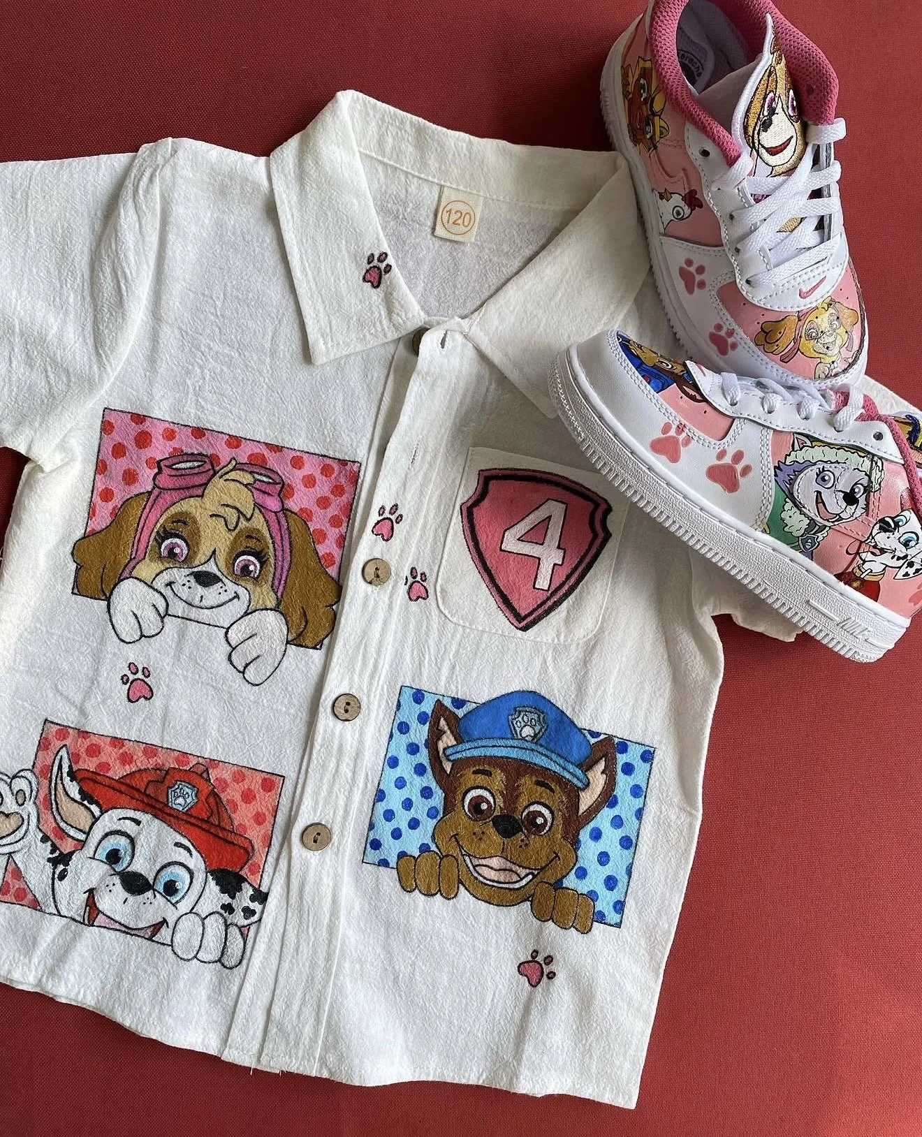 A white shirt with colorful cartoon dog characters, patches, and paw prints, along with a pair of pink and white sneakers featuring cartoon dog designs, all placed on a red surface.