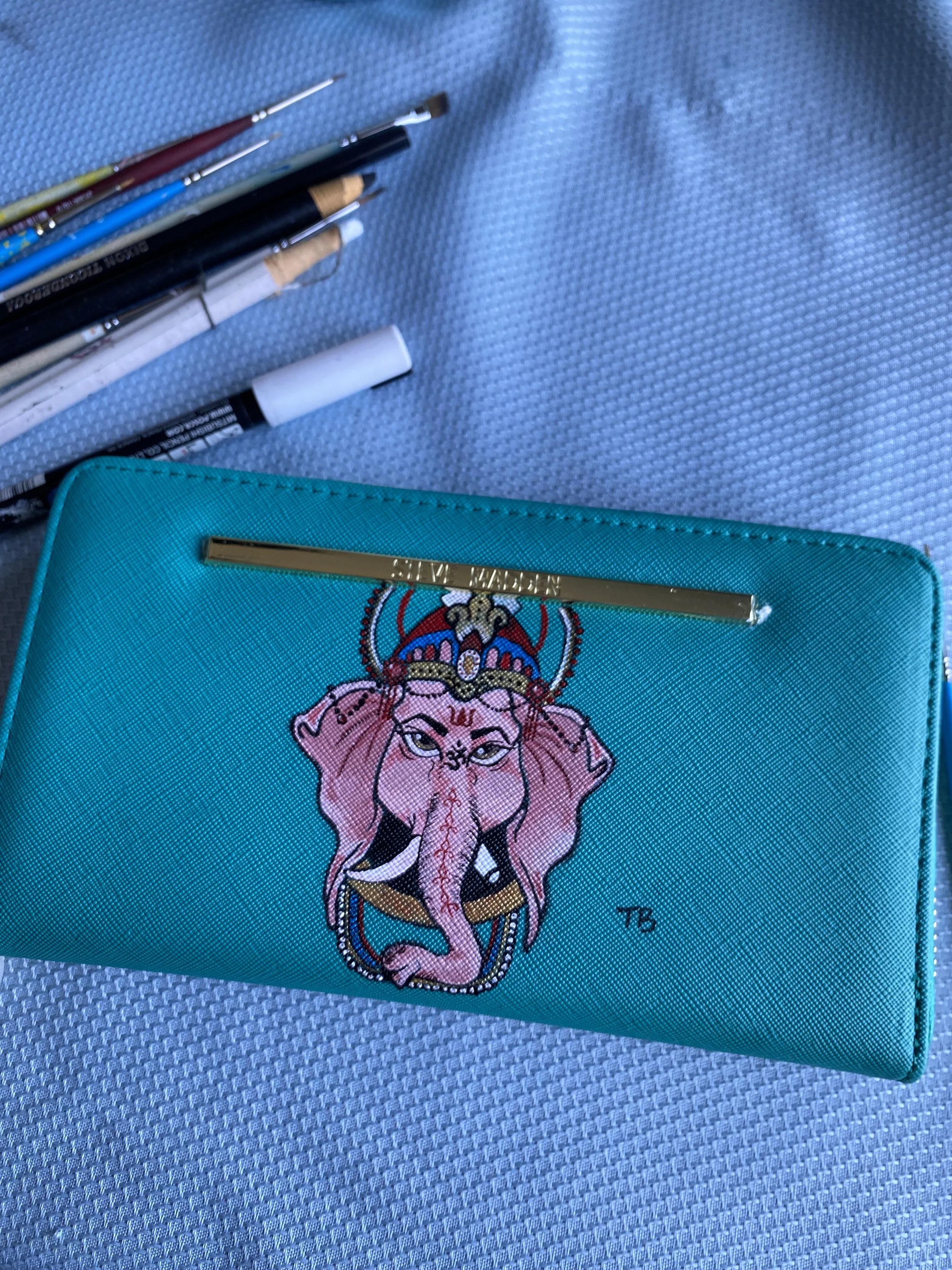 A light blue purse with an embroidered design of an elephant with jewelry on its head and trunk, resting on a textured surface. Several paintbrushes and pens are placed on the surface near the purse.