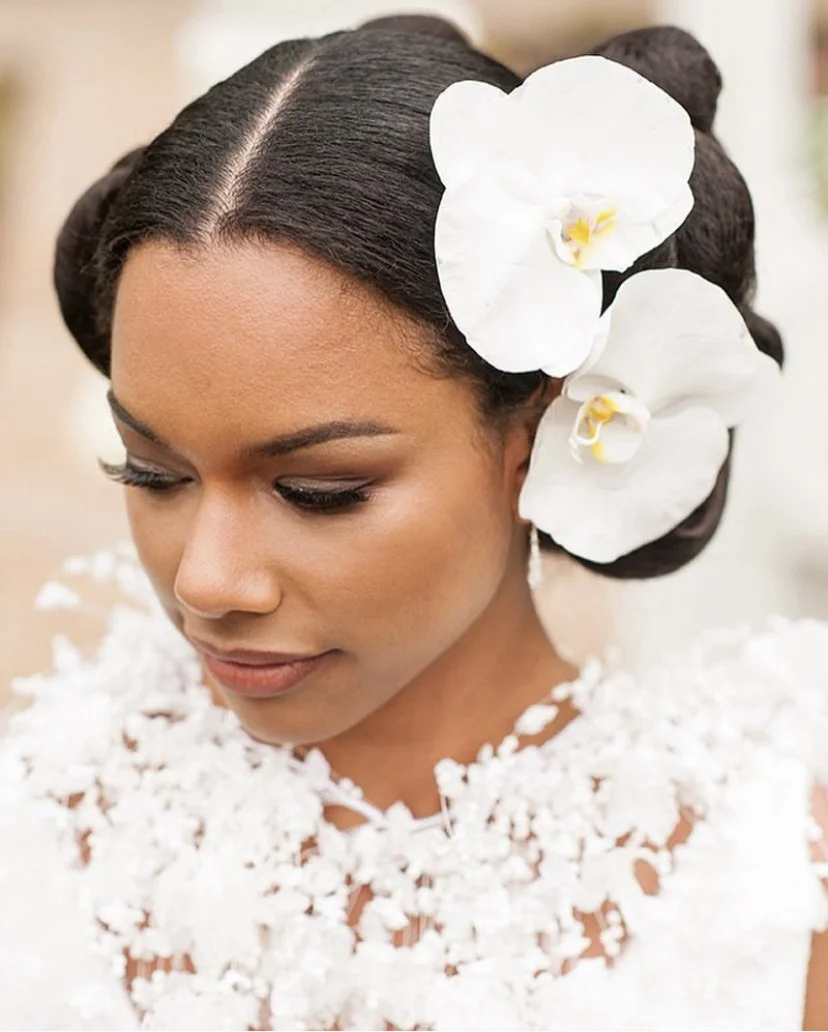 Close-up of a woman with dark hair styled in elegant updo, adorned with white orchids, wearing a white floral dress.