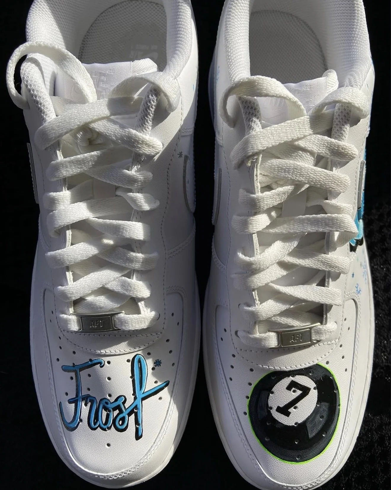 Pair of white sneakers decorated with blue and black marker drawings and writings, placed on a dark surface.