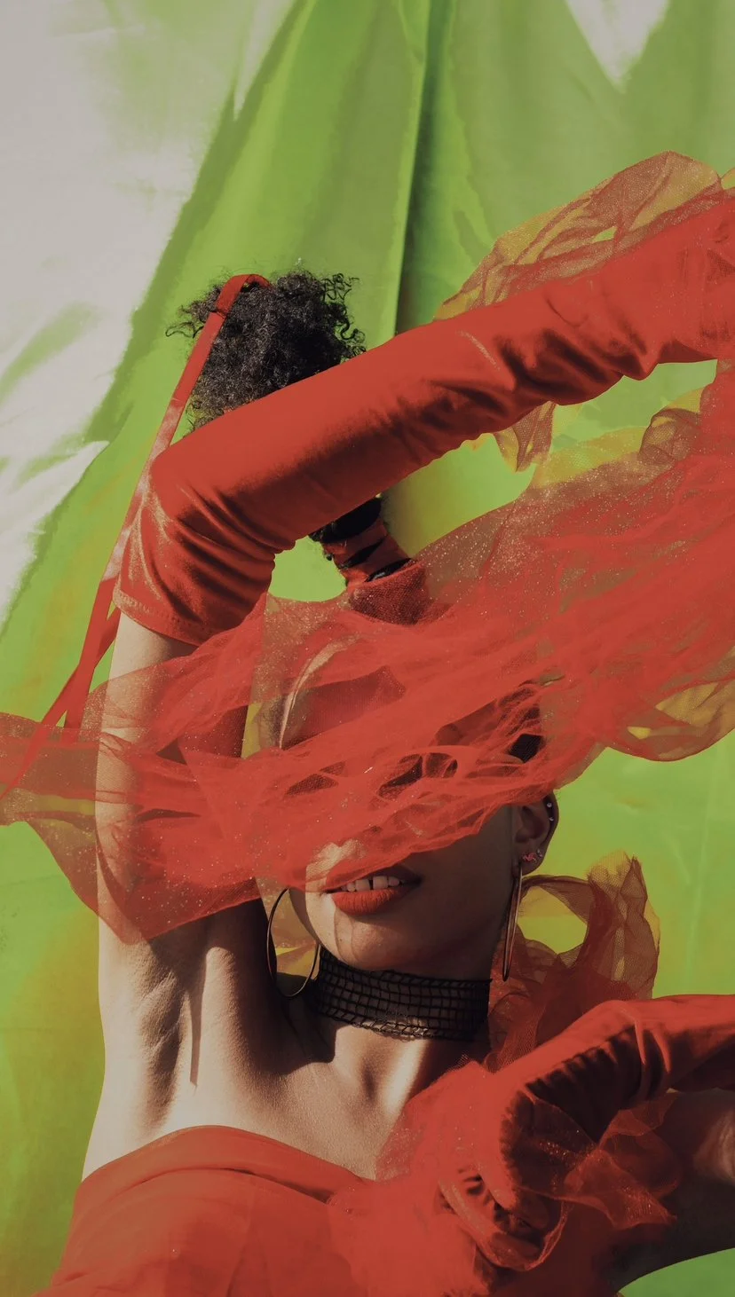 A woman with curly hair is partially covered by orange and red sheer fabric, with a green background and vibrant lighting.