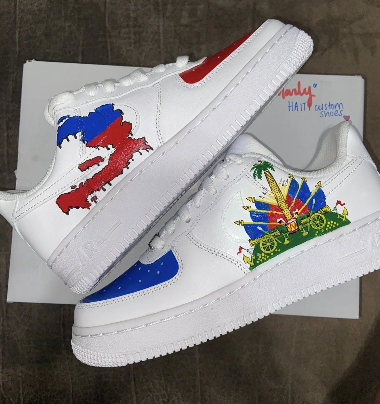 Two custom white sneakers with colorful designs, one featuring a map of Haiti with red, blue, and black paint, and the other depicting a festive scene with a cannon, flags, and palm trees, placed on a shoebox.