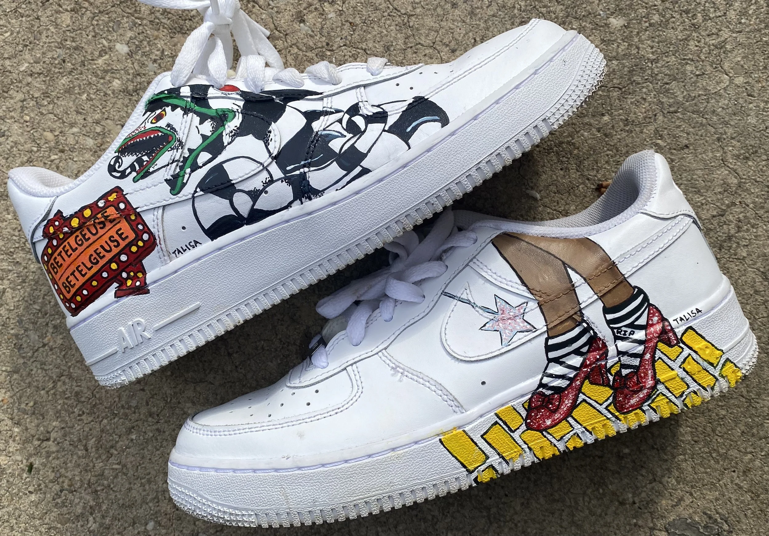 A pair of white sneakers with graffiti-style artwork and colorful designs, placed on a gravel surface.