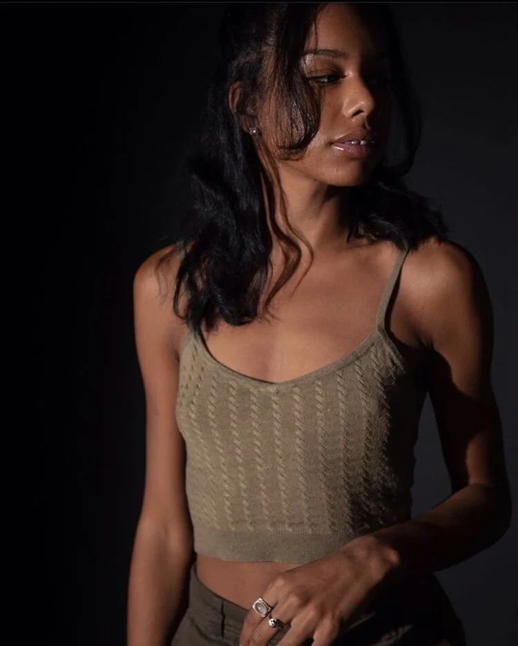 A woman with medium skin tone and dark, wavy hair looking to her right, wearing a beige sleeveless top with thin straps.