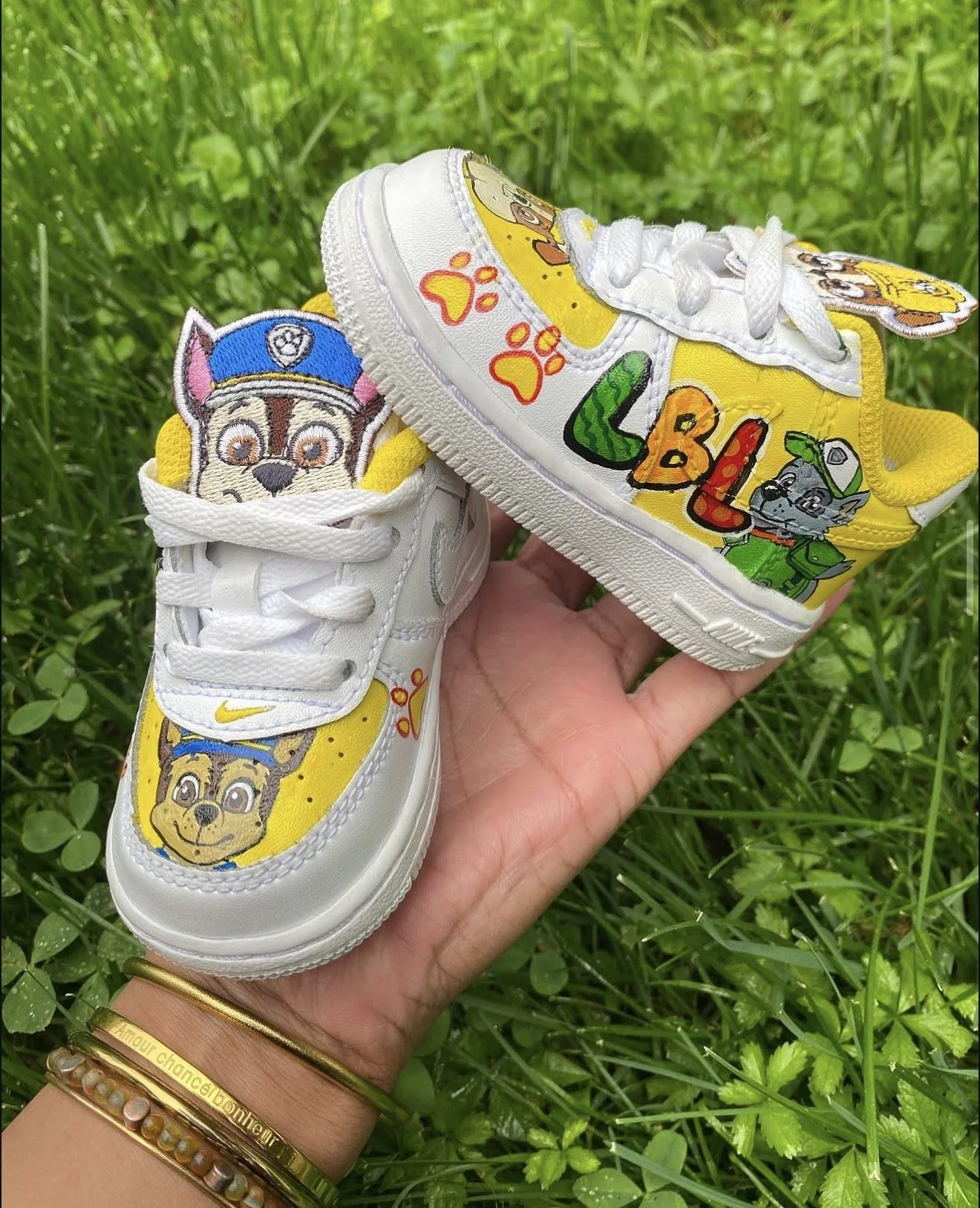 A hand holding a pair of white sneakers decorated with colorful patches of cartoon characters, including Chase and Rubble from Paw Patrol, set against green grass.