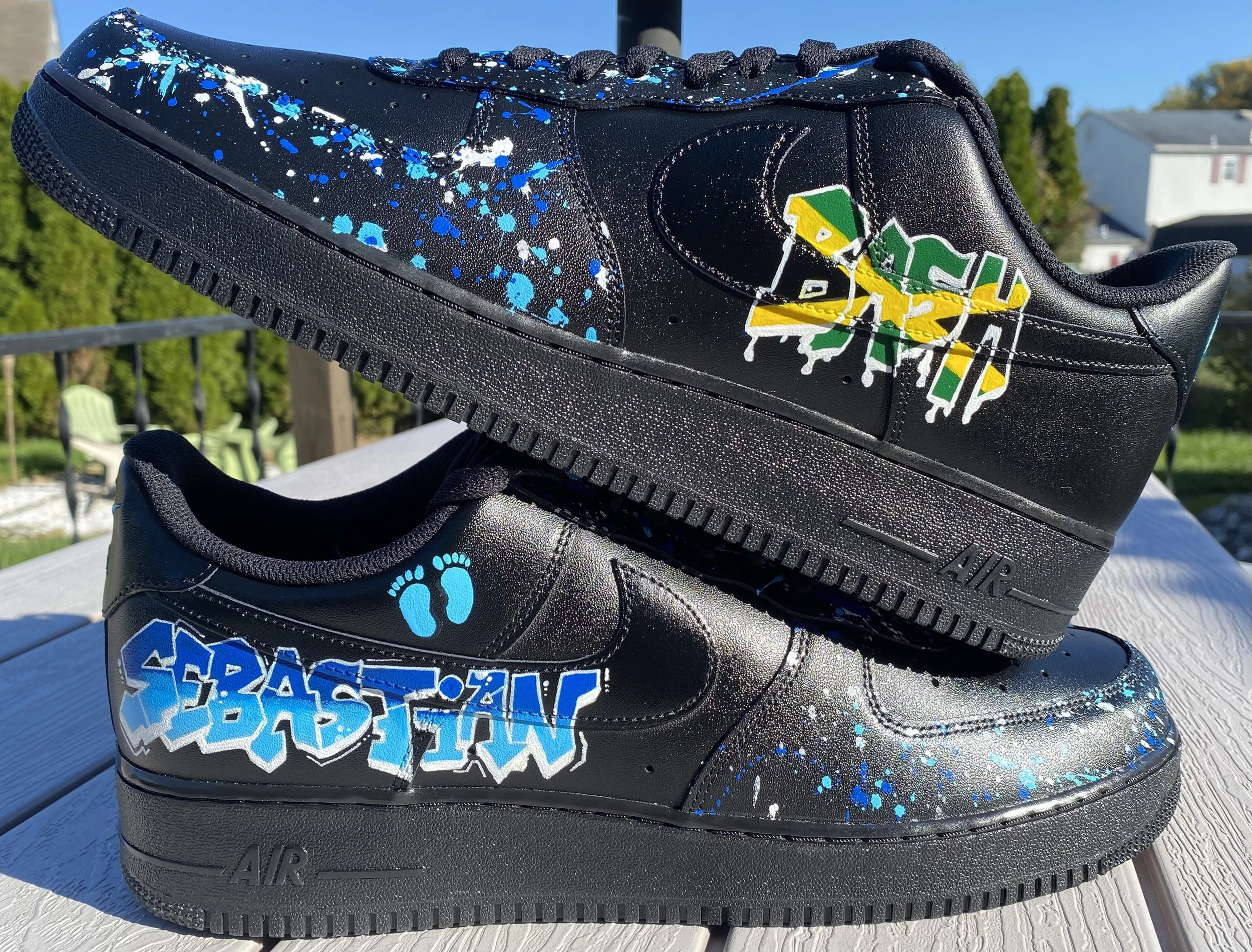Black Nike Air Force 1 sneakers with blue, white, and green paint splatter design and a graffiti-style text sticker, outdoors on a white wooden surface.