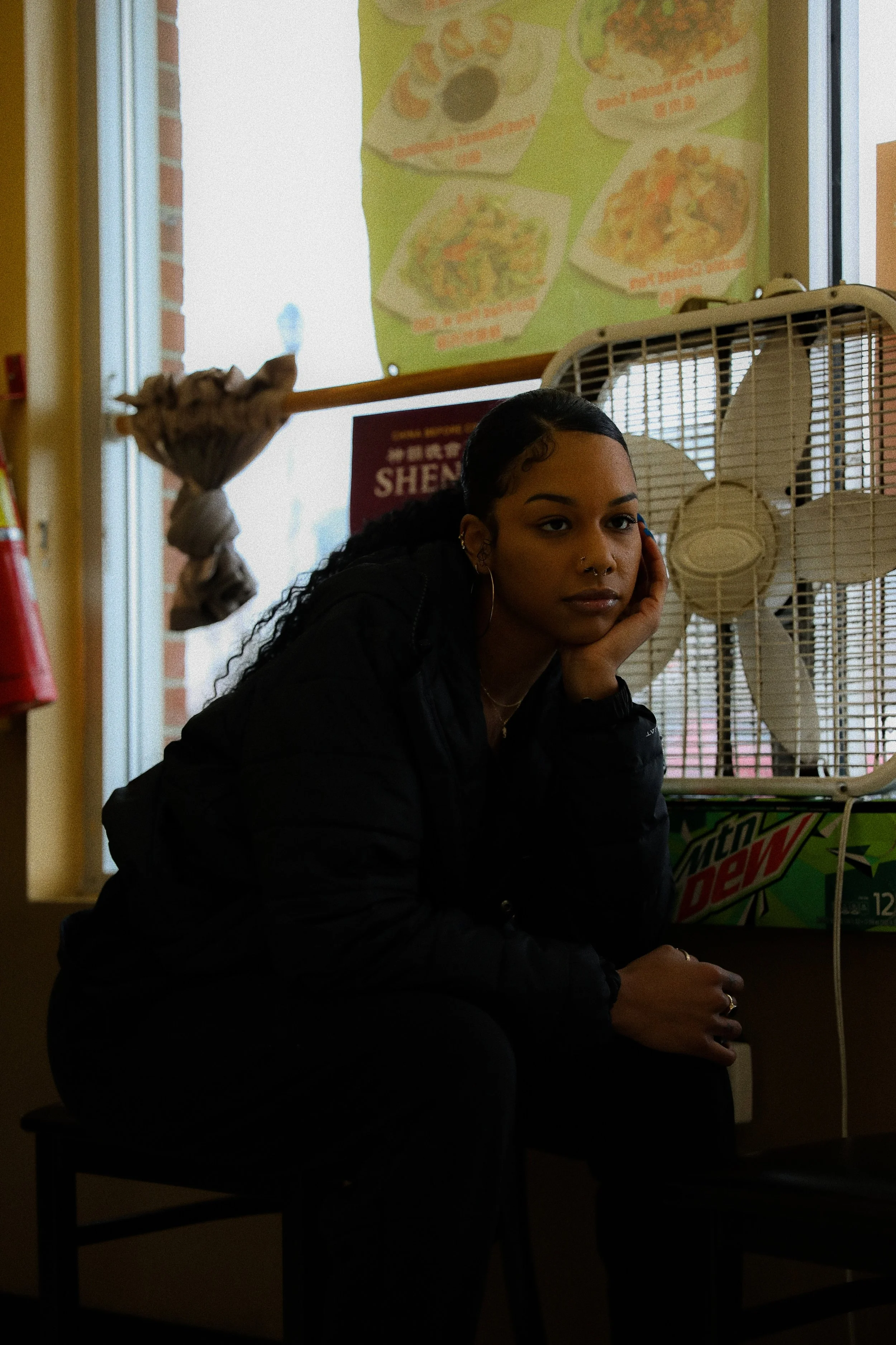 A young woman with dark hair and hoop earrings, sitting at a table, resting her head on her hand near a blonde fan inside a restaurant or convenience store. Moving drink posters are visible in the background.
