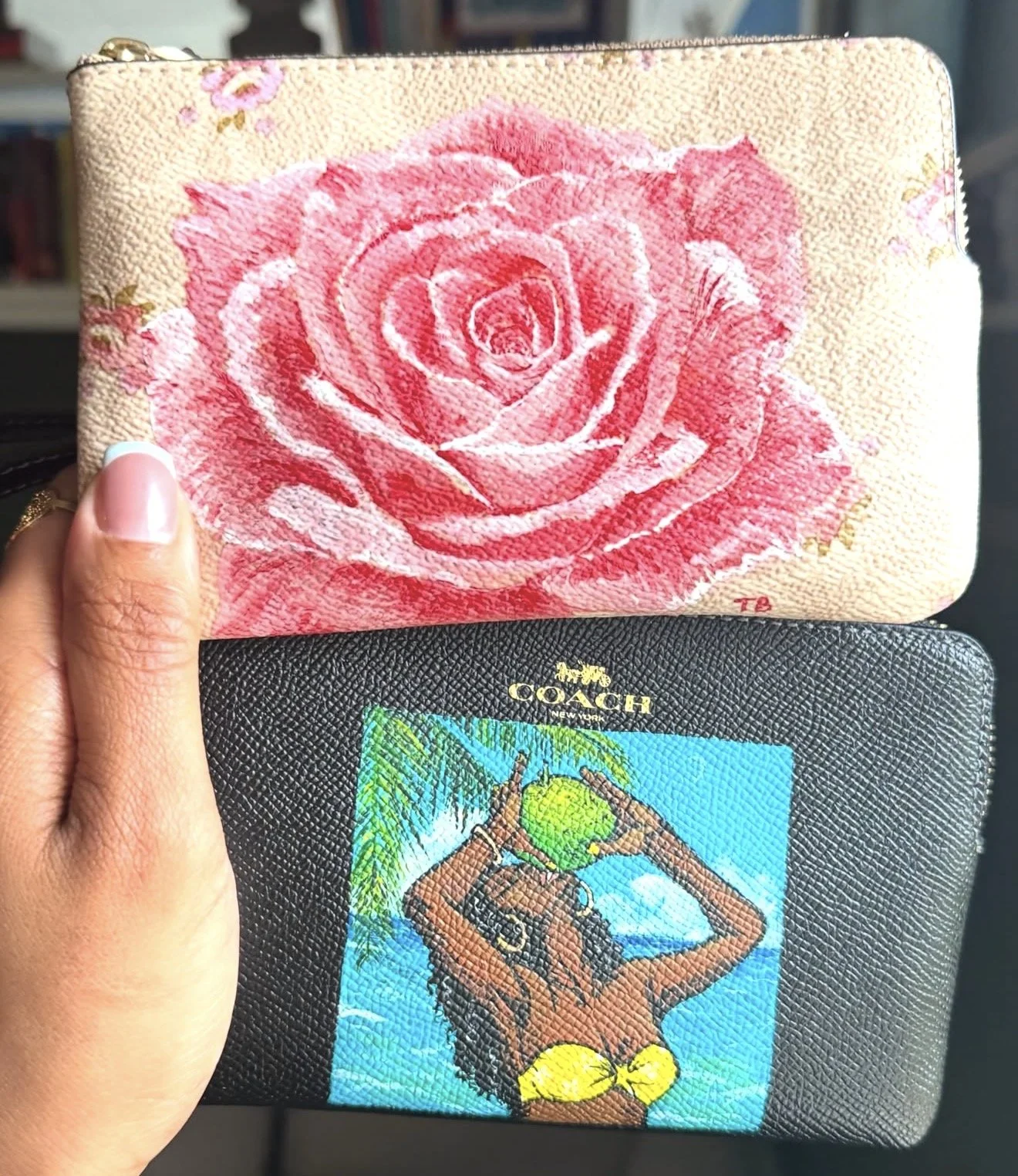 A beige wallet with a pink rose embroidery on the front and a black textured wallet with a depiction of the Girl with a Pearl Earring holding a green sphere.