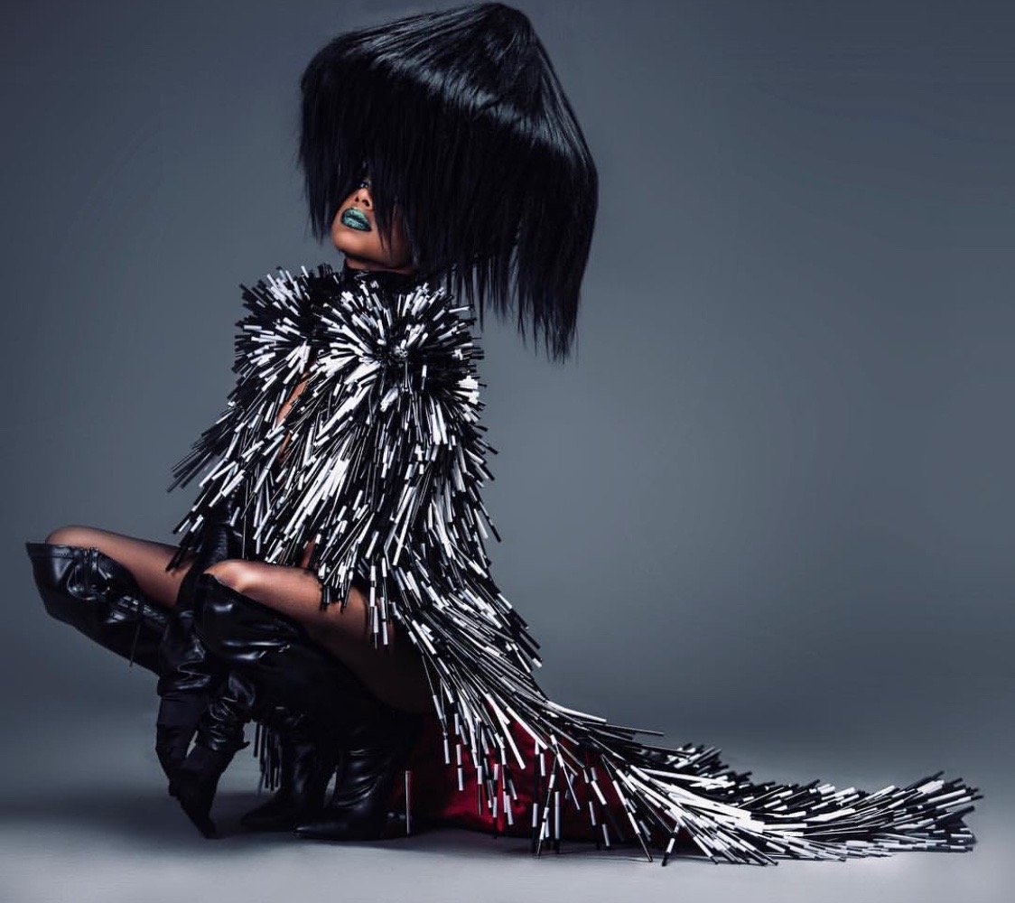 A person with large, black, straight, shoulder-length hair covering their face, wearing a black-and-white spiked dress and black gloves, posing against a gray background.
