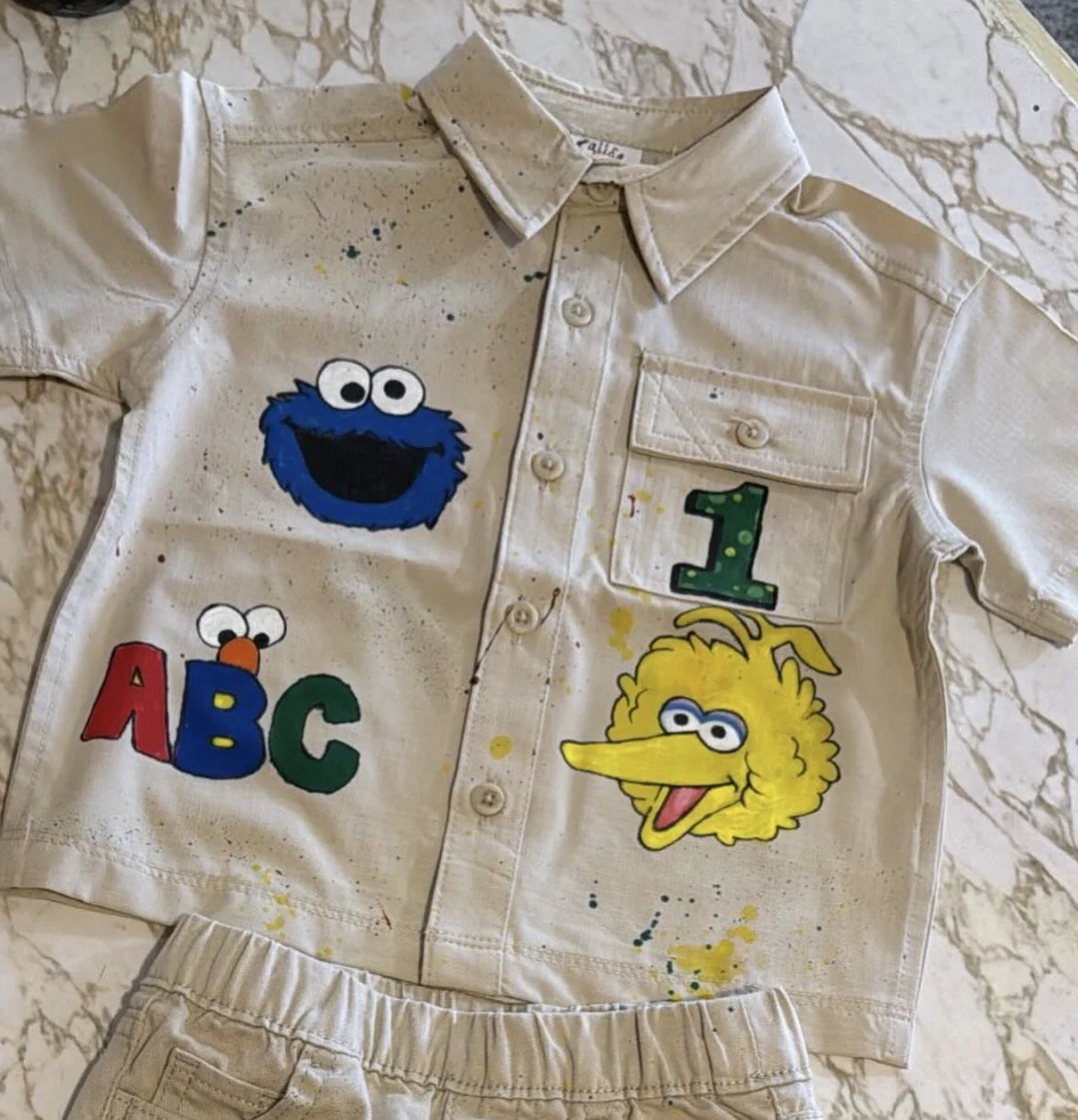 A beige children's shirt and shorts set with Sesame Street character painted designs, including Cookie Monster, Sesame Street logo, Big Bird, and a green number one, with paint splatters.