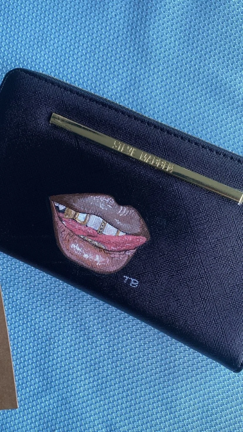 Black clutch purse with a gold bar and the words Steve Madden, featuring an embroidered mouth with lips and gold grills.