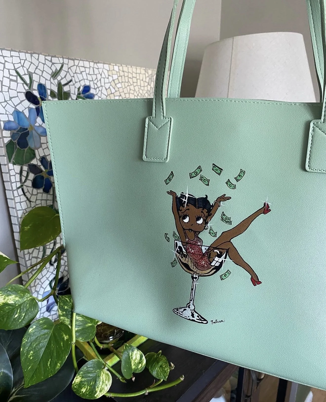 Mint green tote bag with a cartoon illustration of a girl in a cocktail glass, with money flying around, set against a background of assorted houseplants and a mosaic-patterned decoration