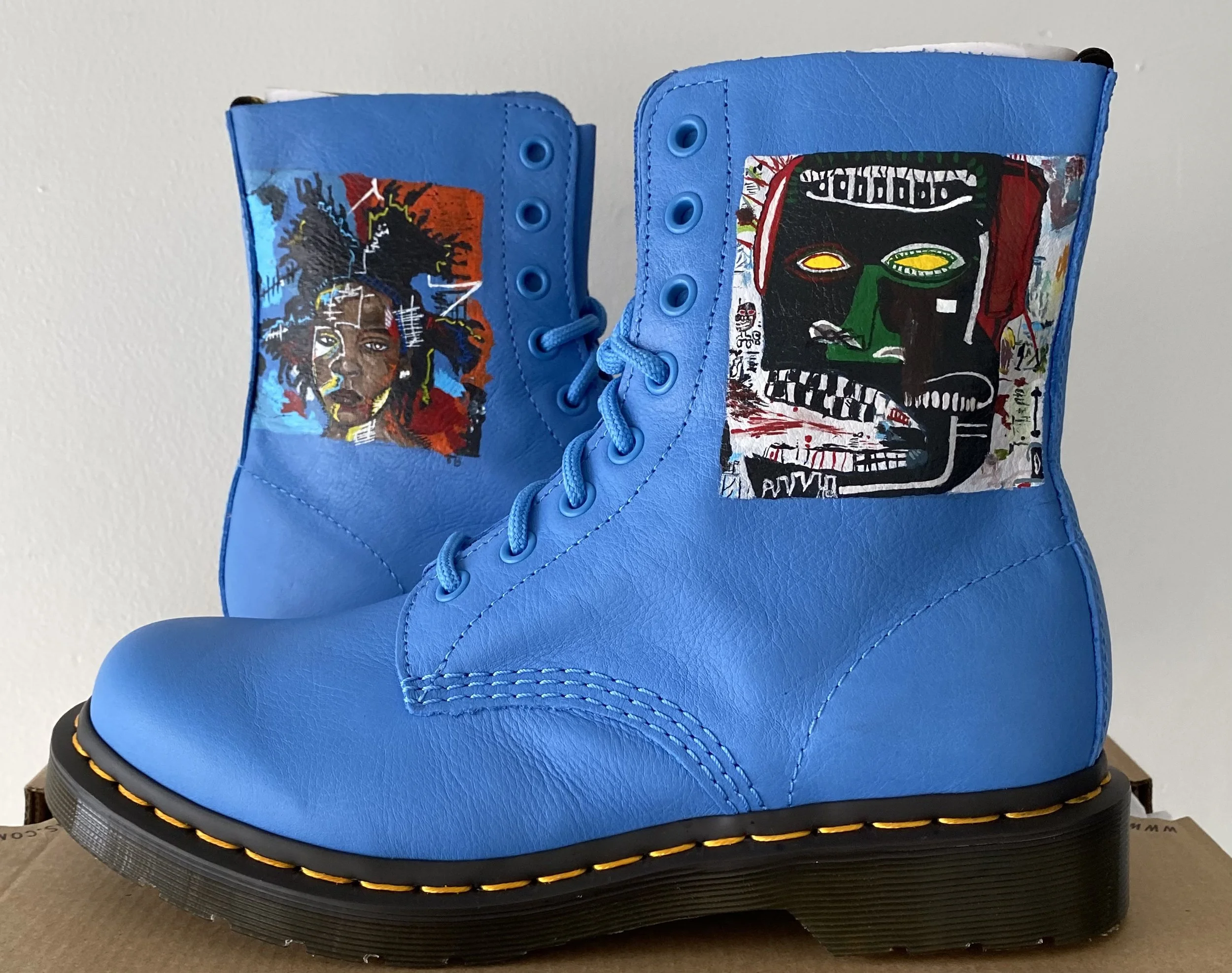 Blue leather lace-up boot with colorful graffiti-style artwork on the side, including faces, abstract shapes, and text, sitting on a black sole with yellow stitching.