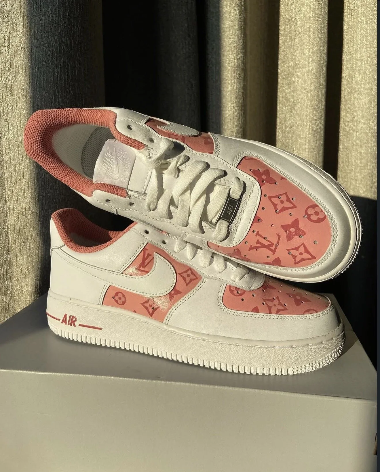 A pair of white Nike Air Jordan sneakers with pink accents and a Louis Vuitton pattern on the leather. The shoes are positioned on a white surface with sunlight illuminating them.