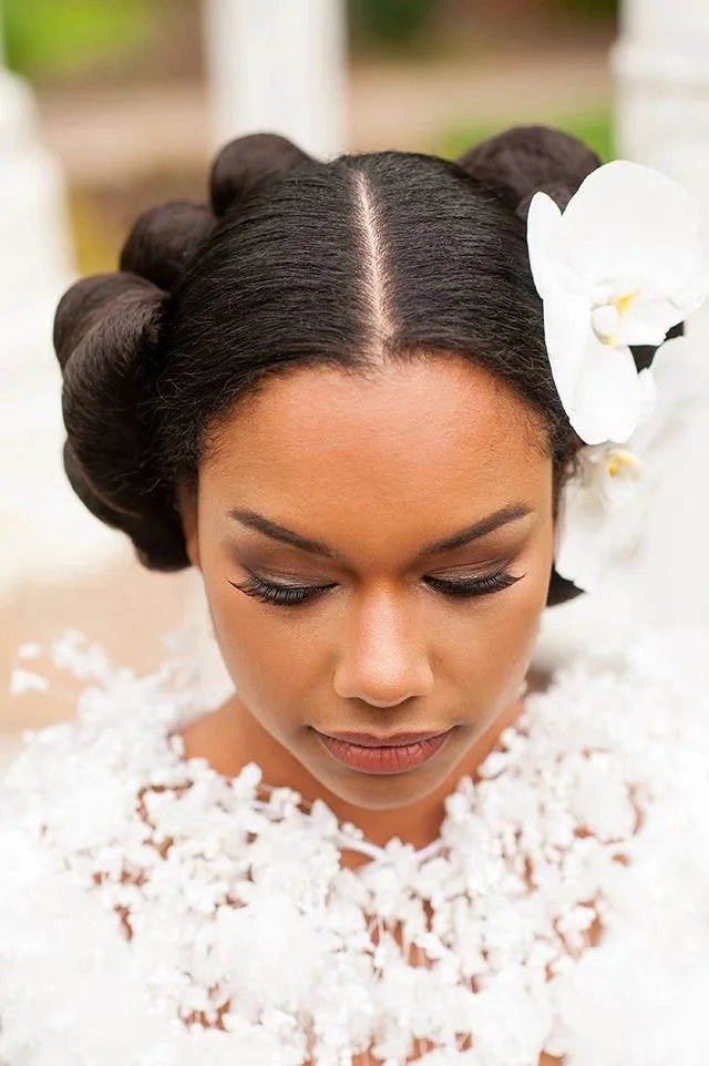 A woman with dark hair styled in vintage rolls, decorated with white orchids, wearing a white floral dress, with eyes closed.