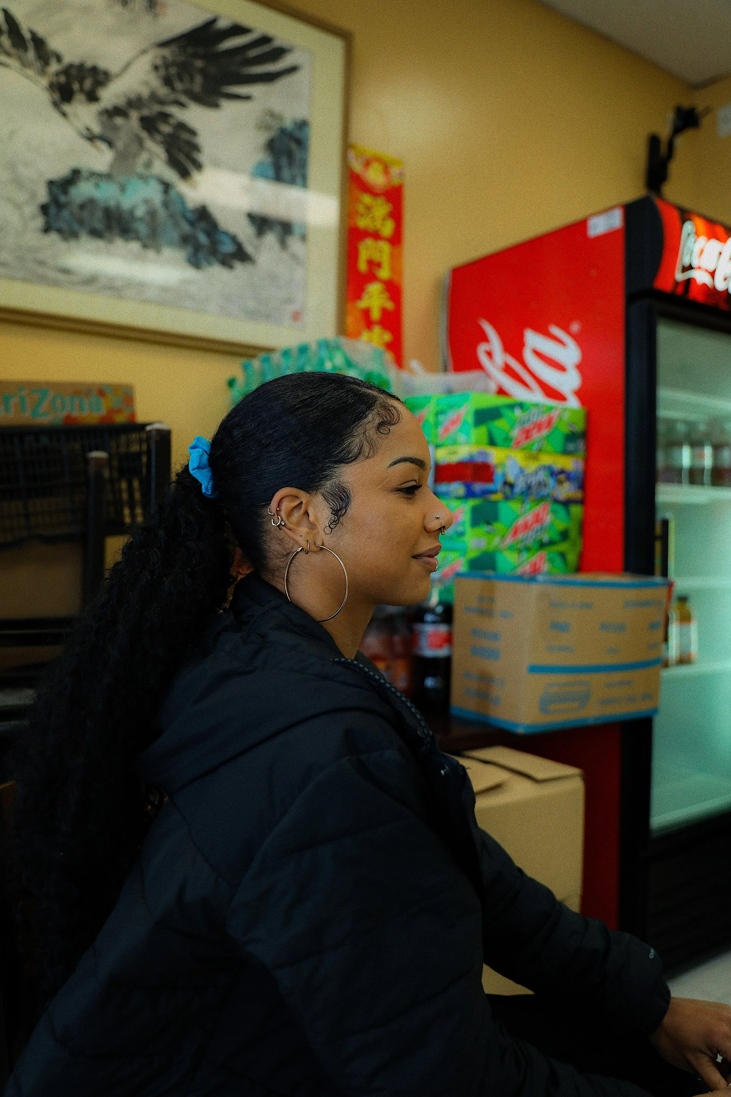 A young woman with curly hair in a ponytail secured with a blue scrunchie, wearing large hoop earrings and a black puffer jacket, is sitting in a convenience store or snack shop. She is facing right, and behind her are stacked boxes of snacks, a vend