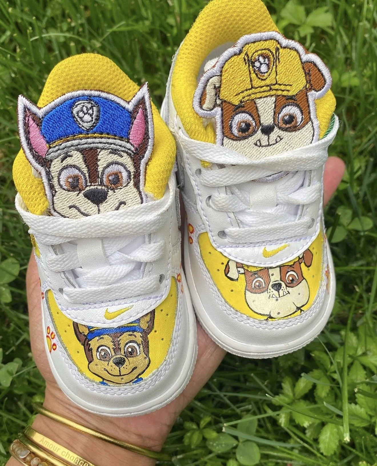 White sneakers with embroidered dog characters on the tongue and toe area, featuring a police dog on one shoe and a firefighter dog on the other, against a grassy background.