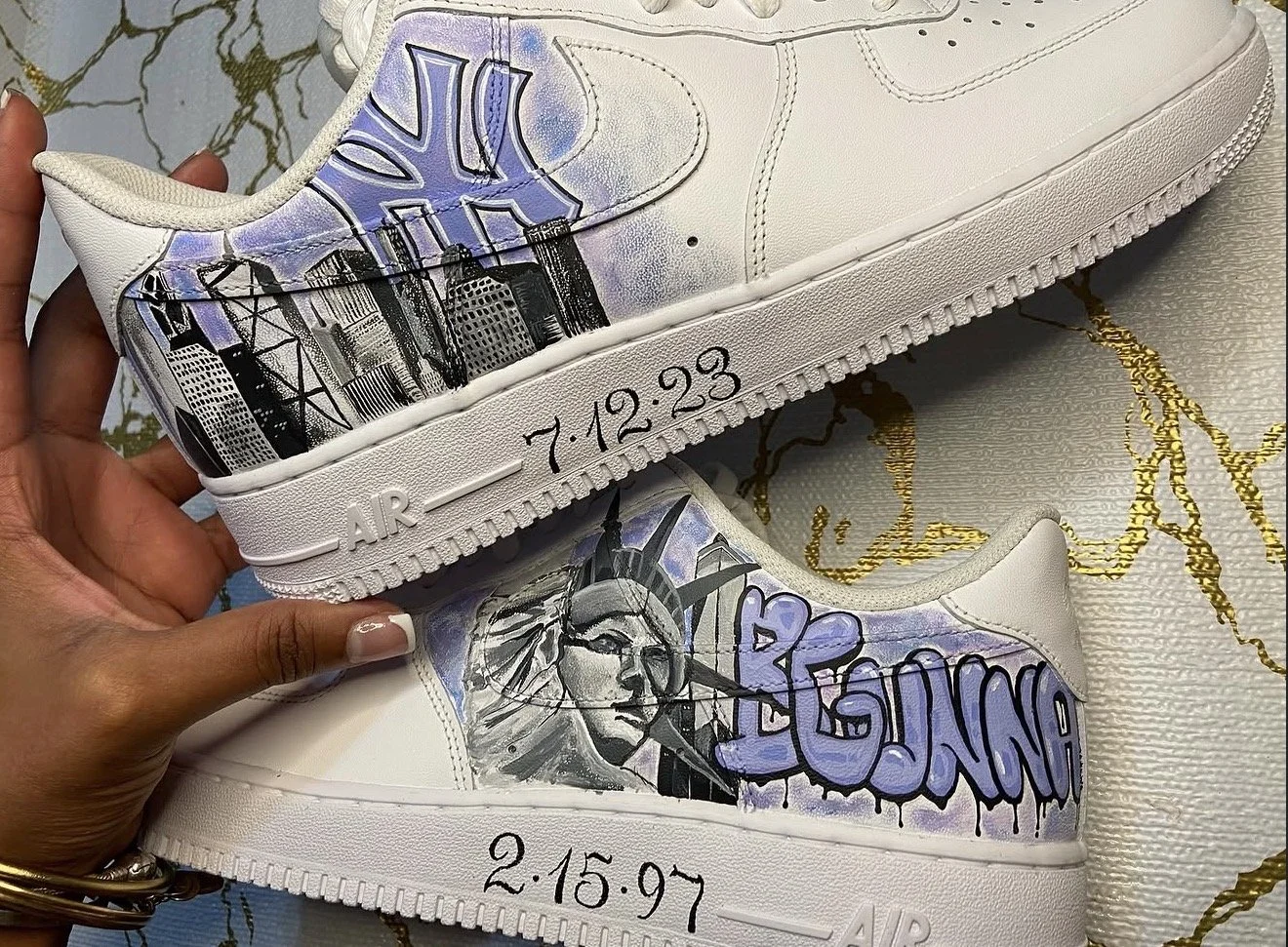 Pair of white sneakers with black and purple graffiti-style art depicting a cityscape and a woman's face, along with dates 7.12.23 and 2.15.97.