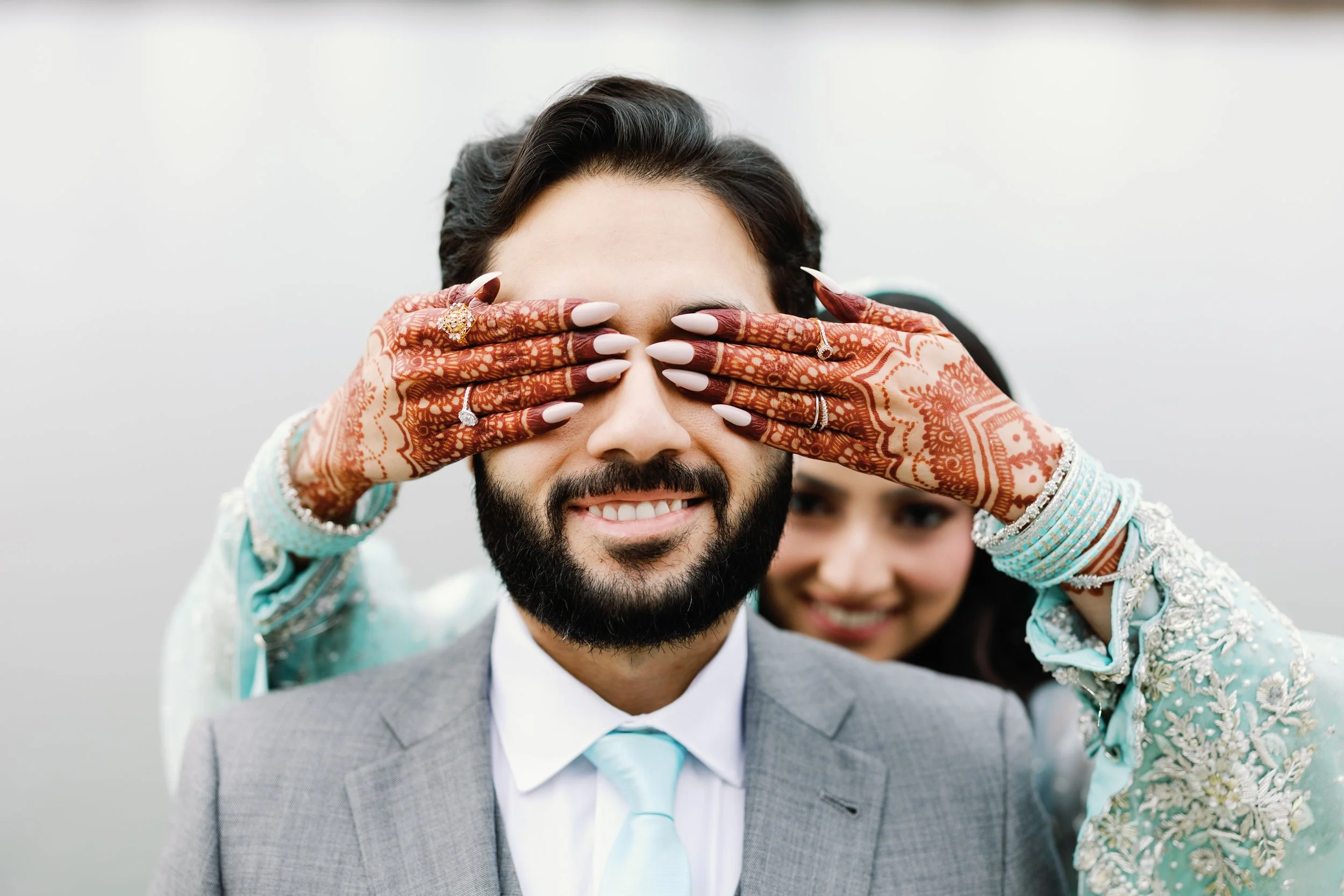A groom dressed in a grey suit with a blue tie, smiling, is being playfully covered by a bride's hands with intricate henna designs on her fingers, and wearing rings. The bride's face is partially visible behind him, also smiling. The background is p
