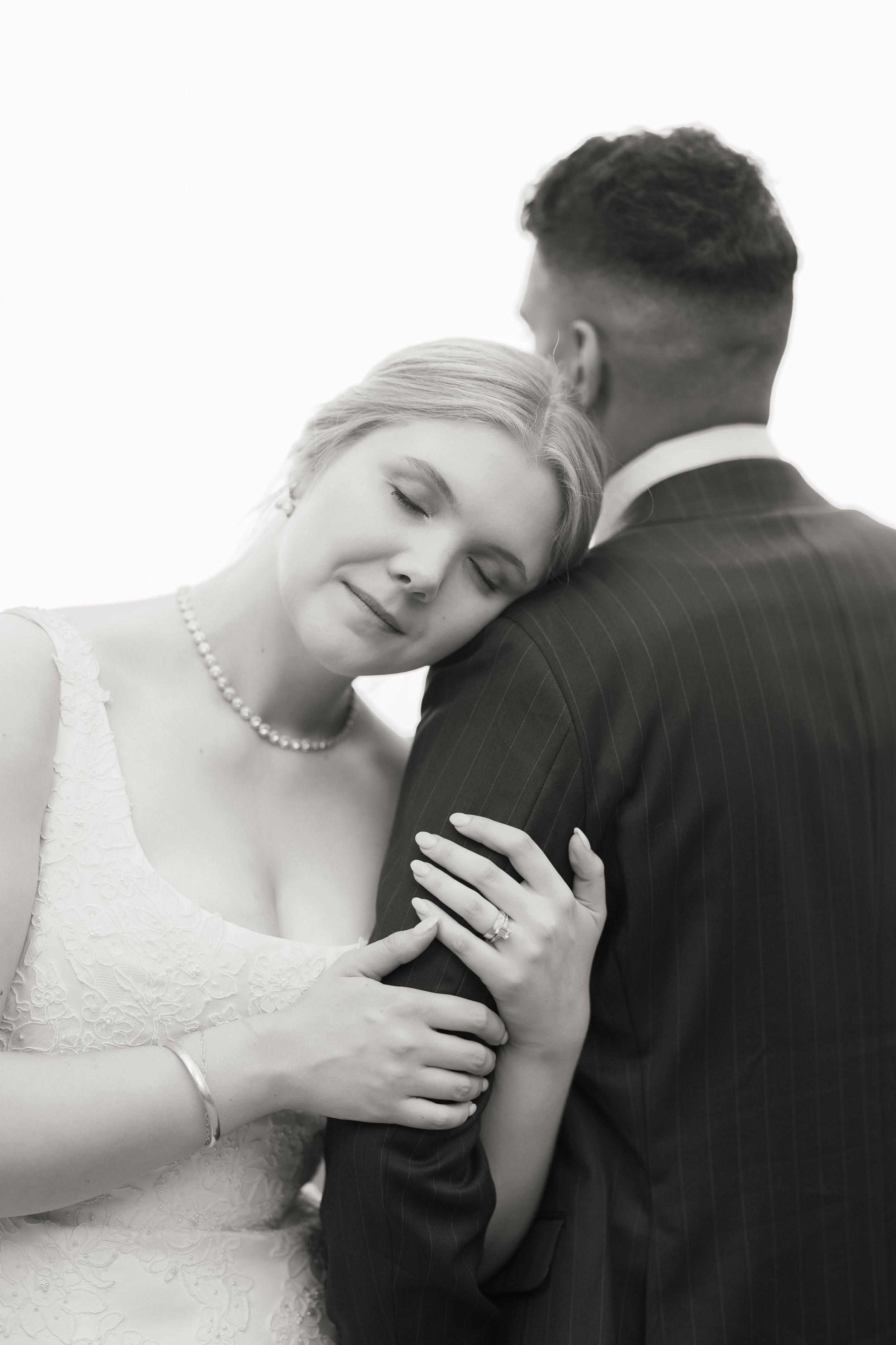 A black and white photograph of a bride and groom, with the bride resting her head on the groom's shoulder and eyes closed. The bride wears a lace dress, pearl necklace, and ring, while the groom wears a pinstripe suit.
