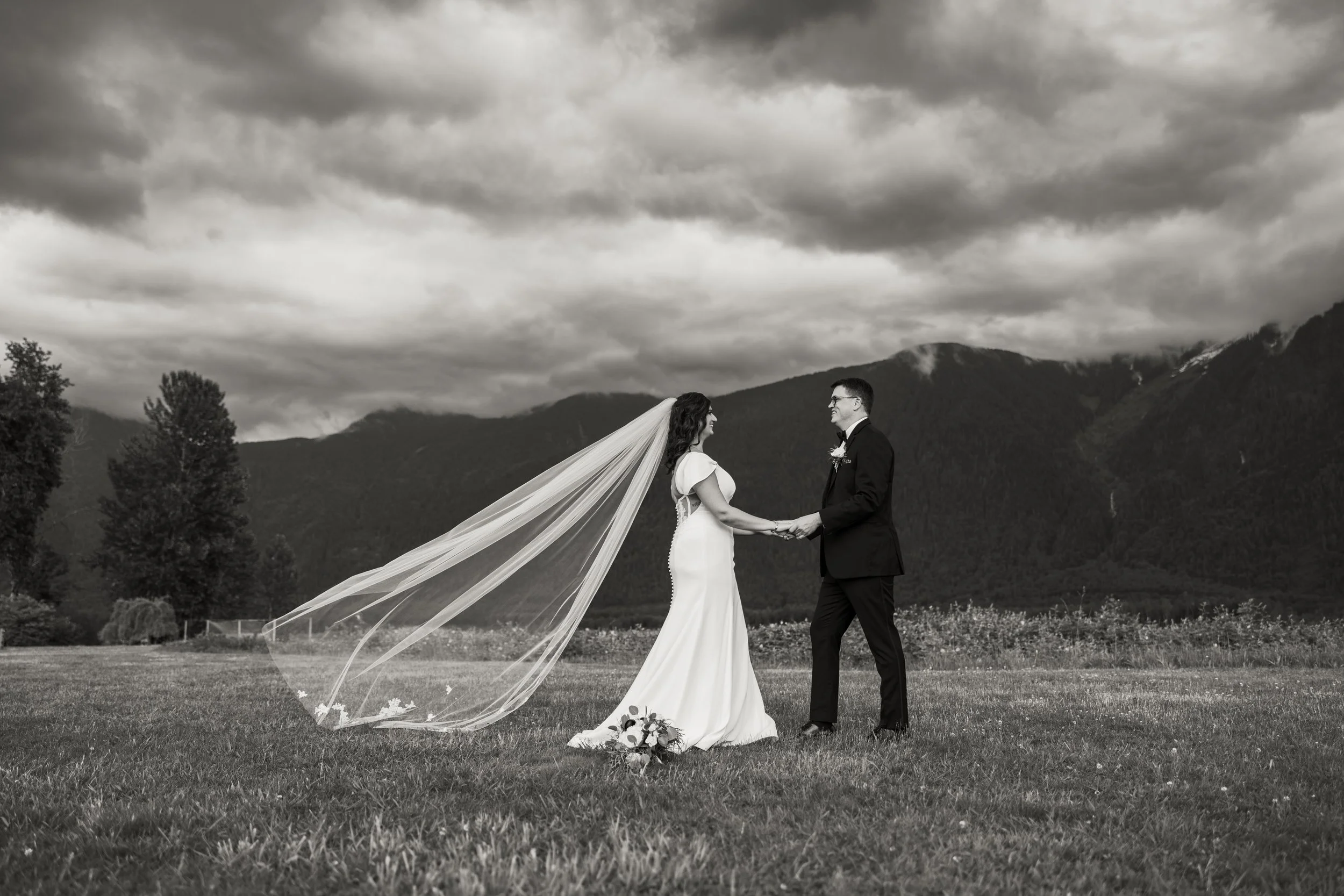 Black-and-white photo of a bride and groom holding hands outdoors at Fraser River Lodge with mountains and cloudy sky in the background.