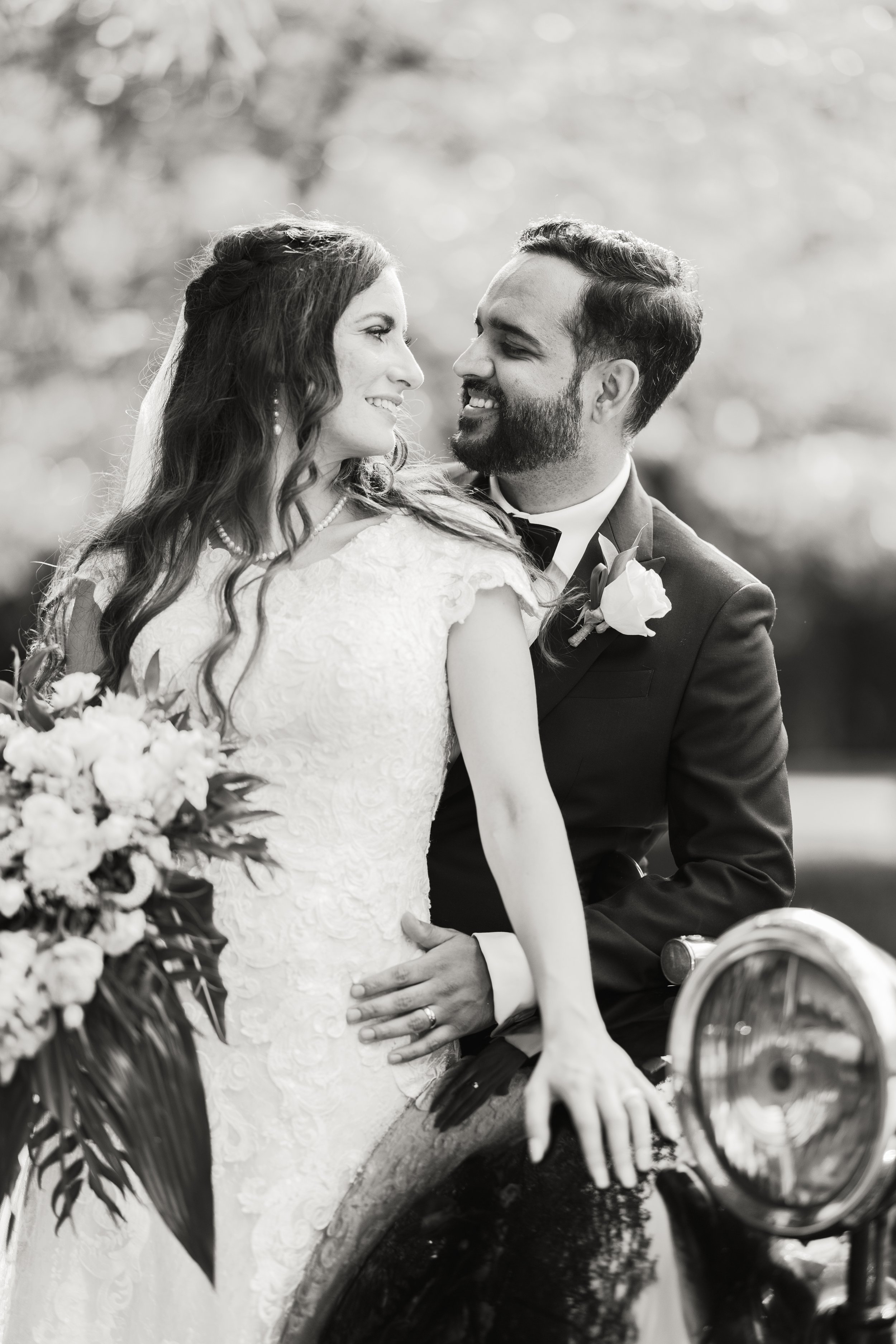 Black-and-white photo of a bride and groom in wedding attire, looking at each other affectionately, with the bride holding a bouquet and sitting on a vintage motorcycle.