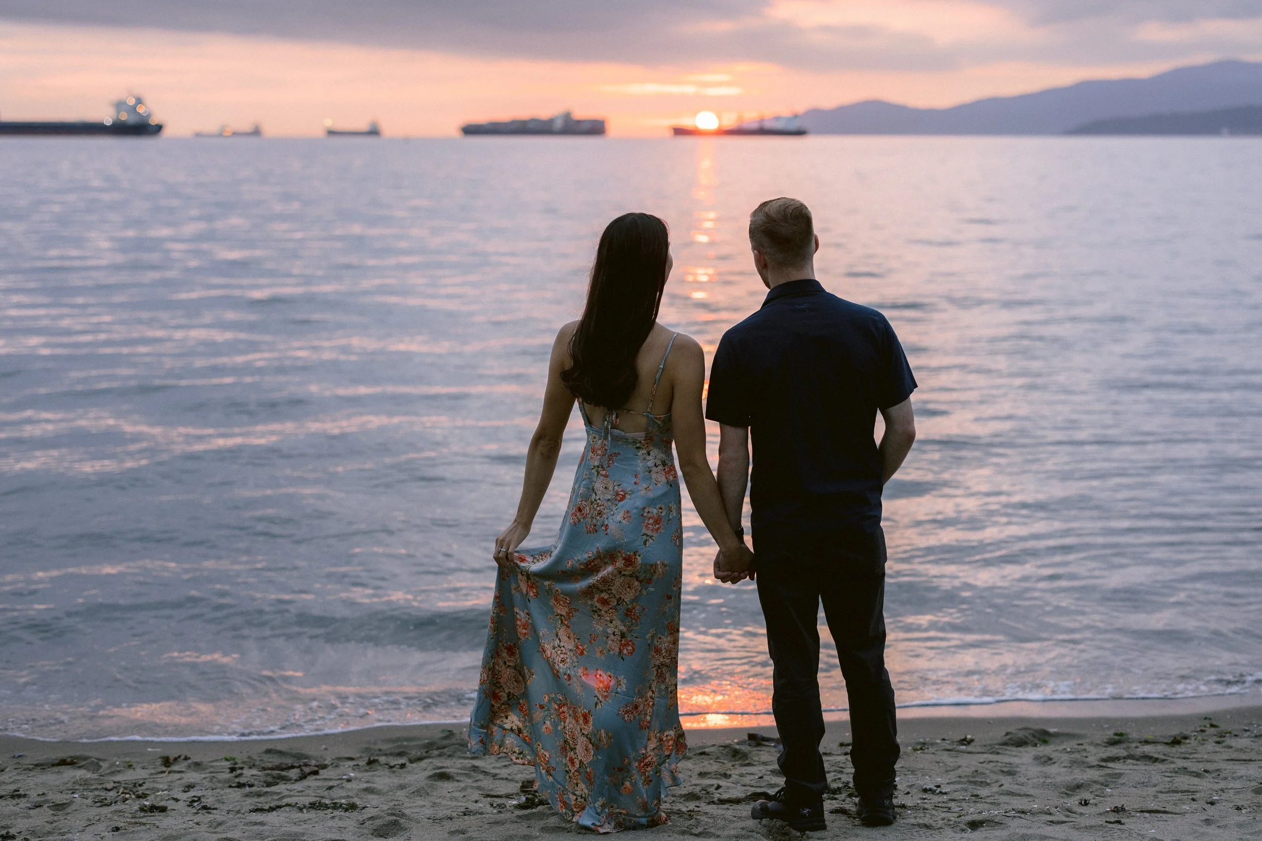 An engaged couple holding hands and walking along the beach at sunset, with ships on the water in the background.