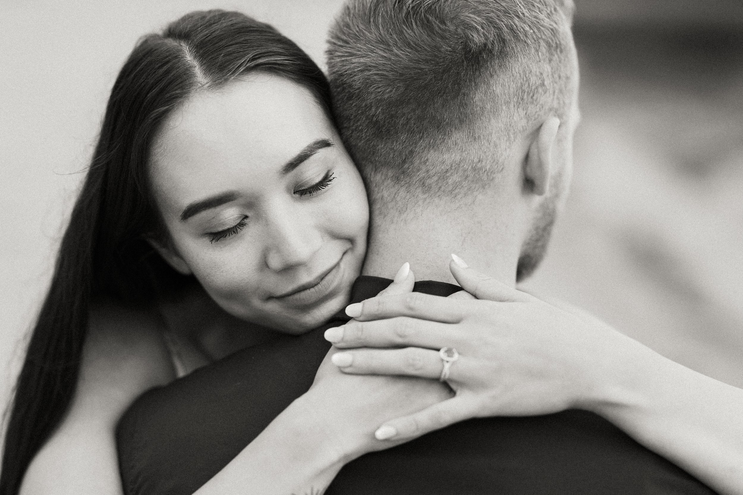 A black-and-white photo of a woman with closed eyes hugging a man, with her head resting on his shoulder. Her hands are gently placed on his shoulder.