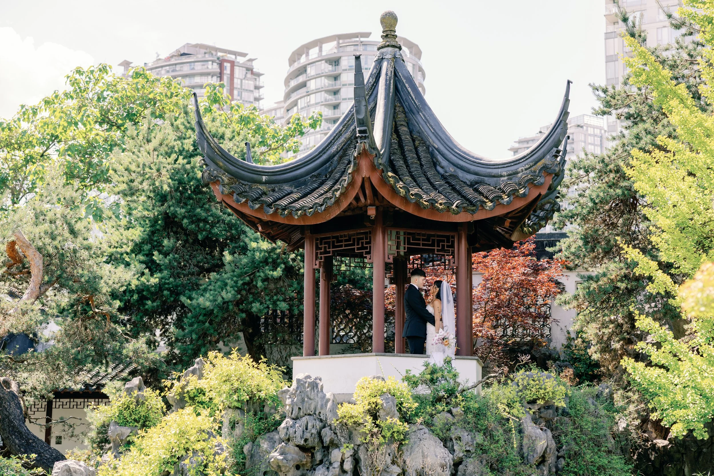 A couple dressed in wedding attire, holding hands, standing inside a traditional Chinese pavilion surrounded by lush greenery in Dr. Sun Yat-Sen Classical Chinese Garden.
