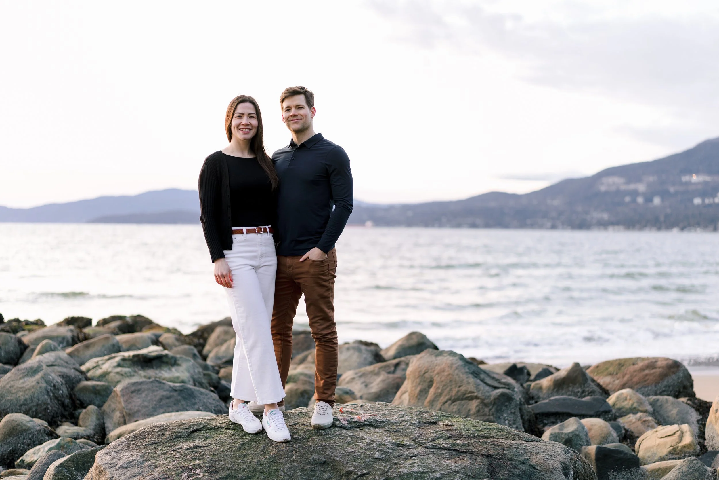 A smiling engaged couple standing on rocks along a body of water with mountains in the background during daytime.