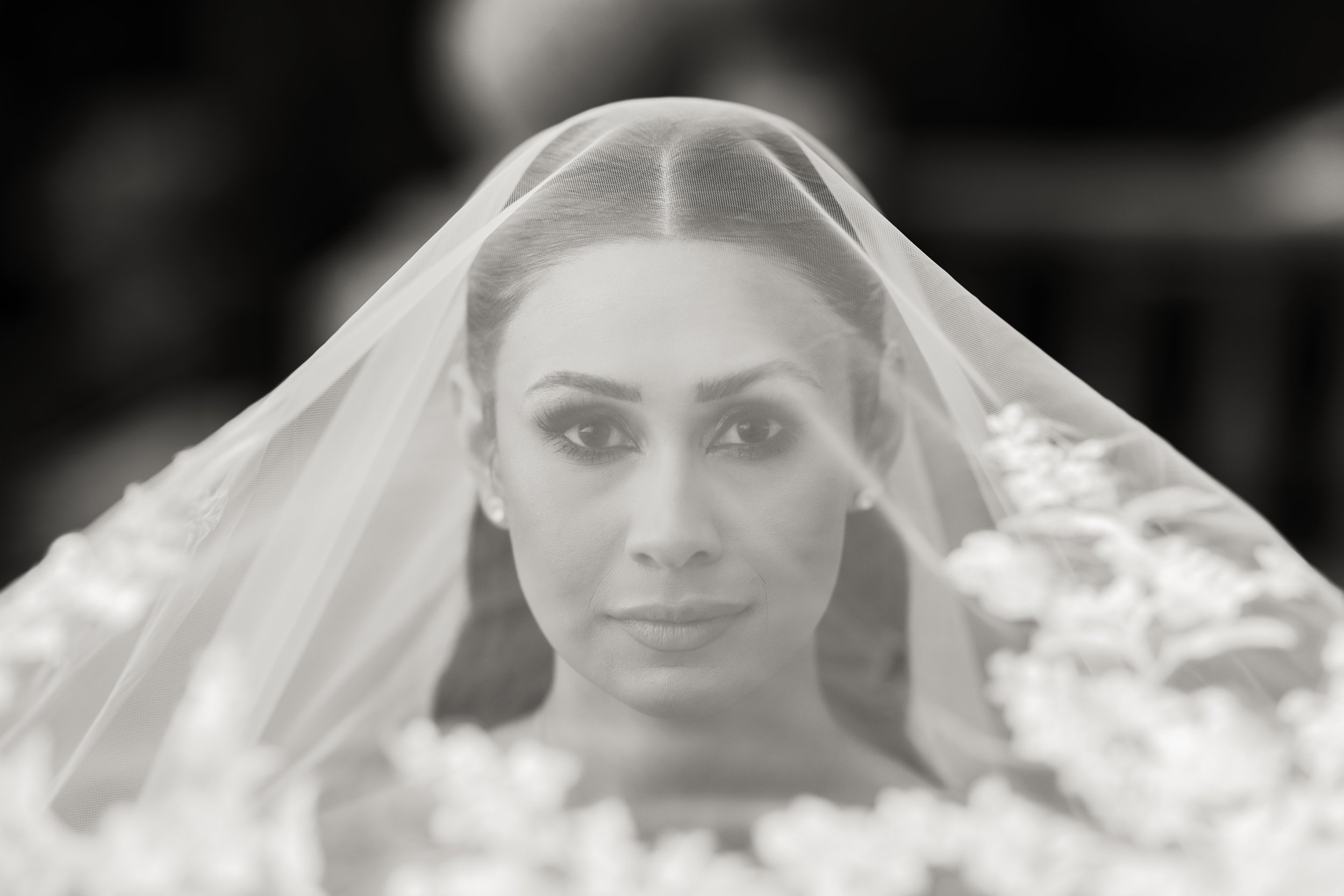 Black and white portrait of a bride with a veil covering her head, looking directly at the camera.