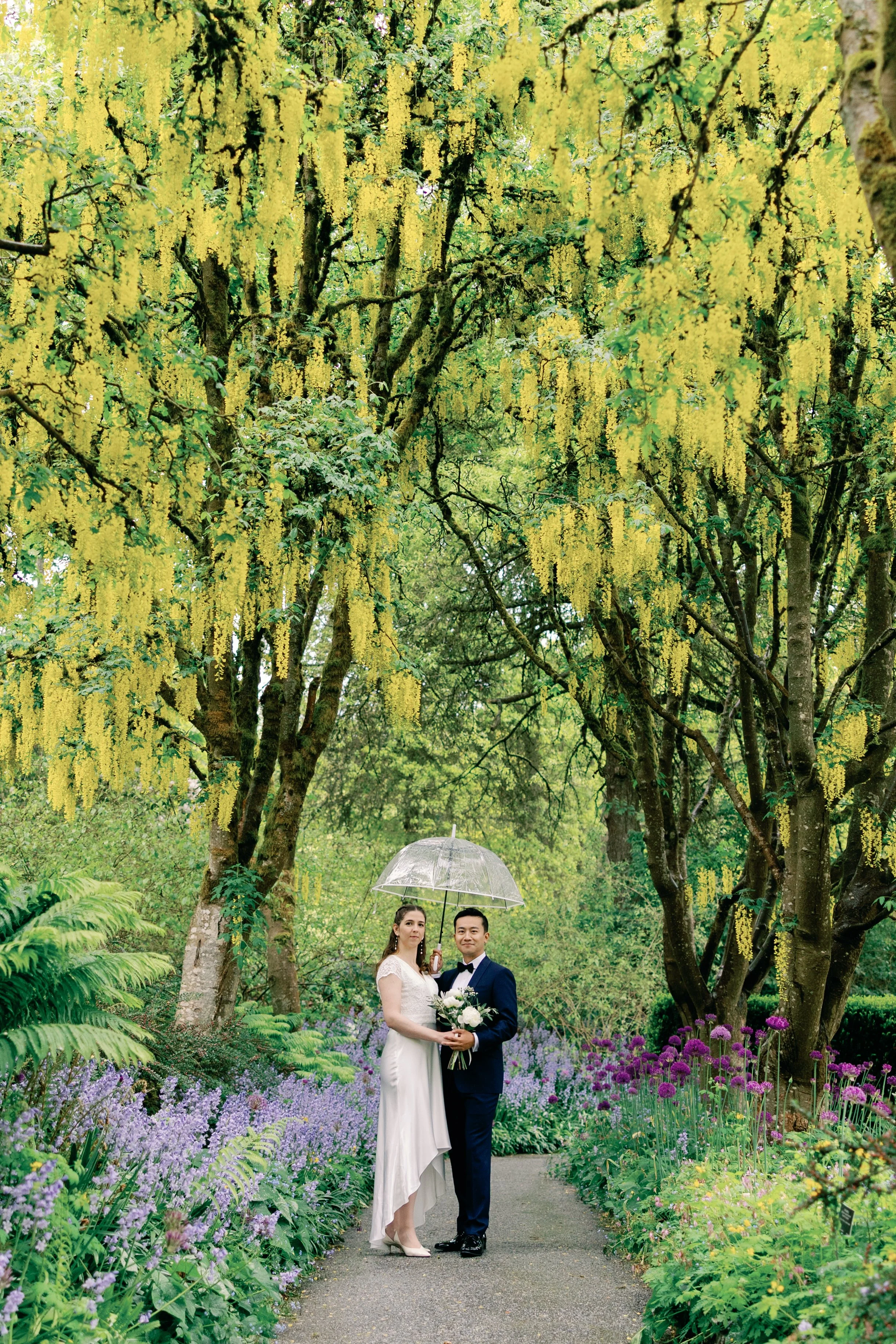 A bride and groom standing together on a garden path during a rainy day, with the bride holding a transparent umbrella and a bouquet of white flowers, surrounded by lush green foliage and flowering plants in Vandusen Park.