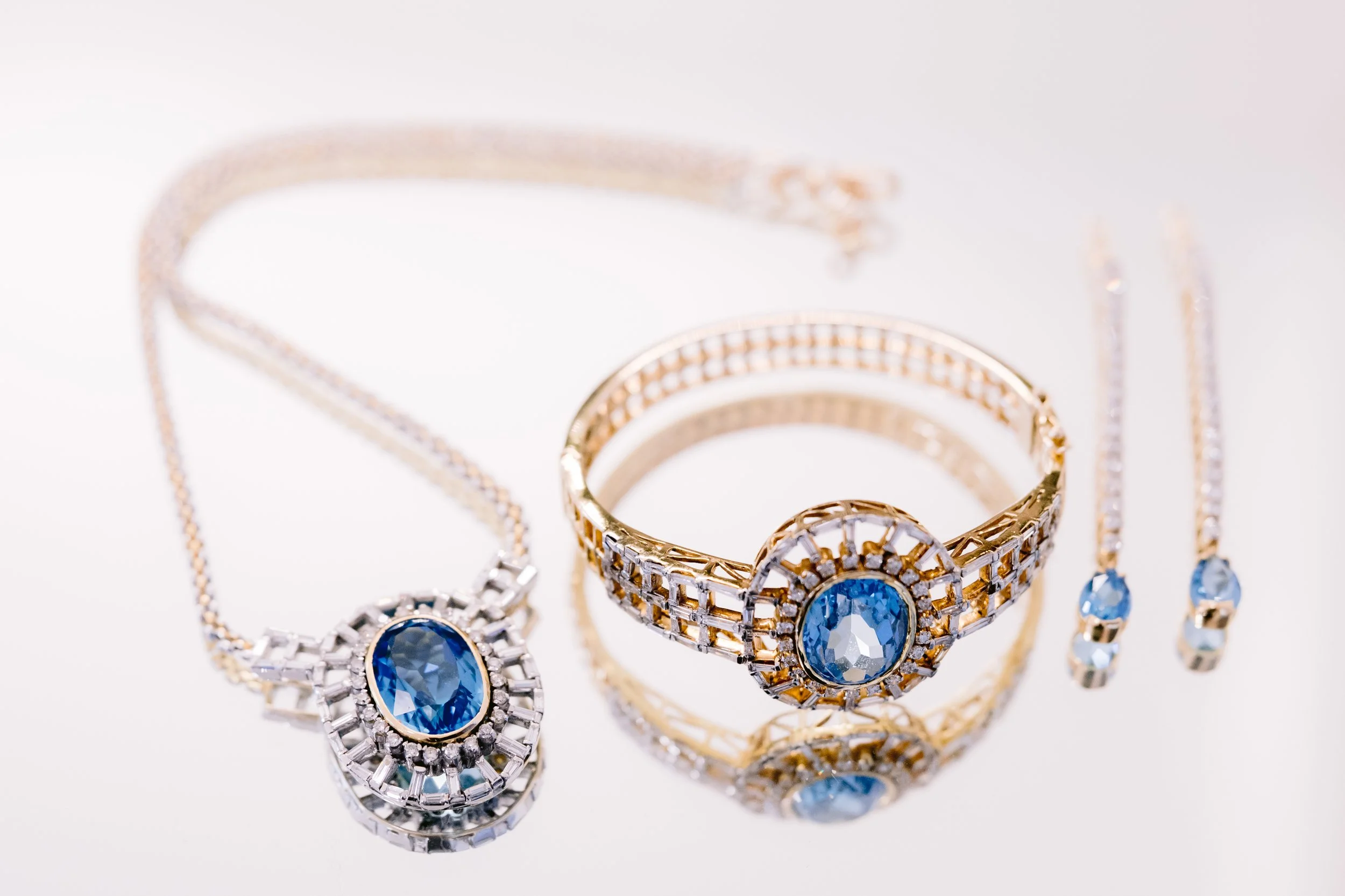 Gold and silver jewelry set with blue gemstones, including a necklace, bracelet, and earrings, on a reflective white surface.