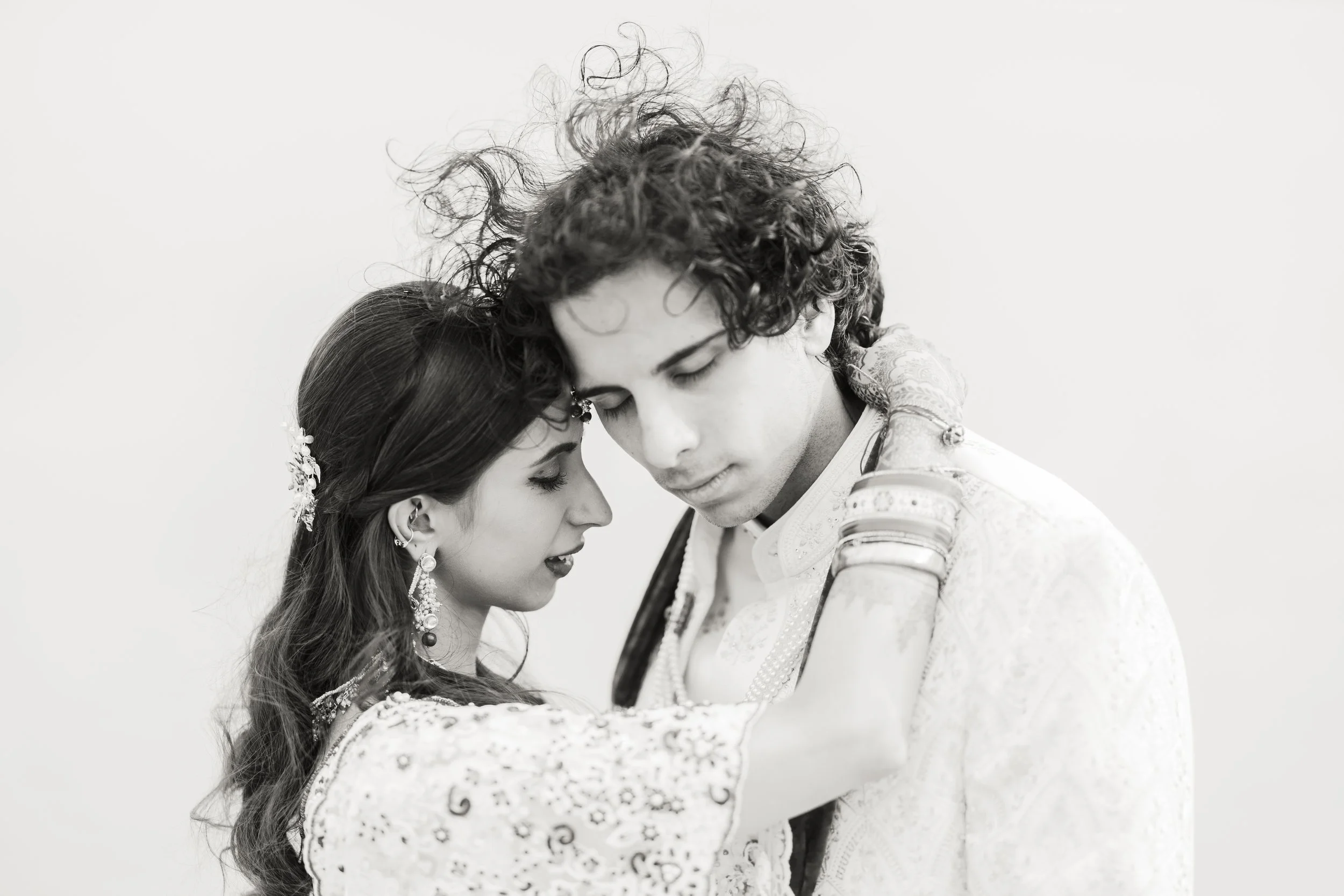 A black and white photo of a couple embracing closely, with their foreheads touching. The bride has long hair and jewelry, and the groom has curly hair and wears traditional Indian attire.