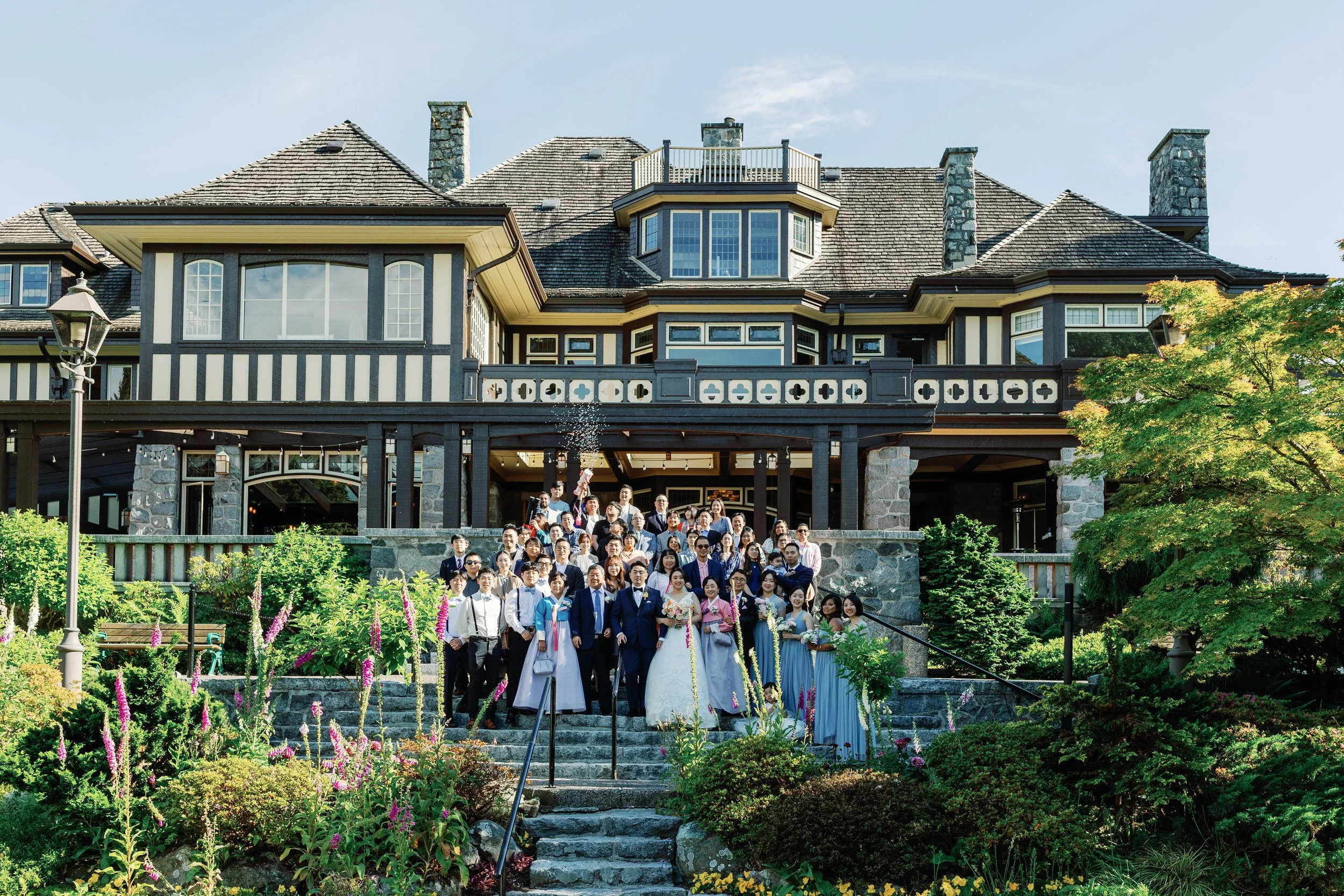 Wedding guests dressed in formal attire gathered on the steps and lawn in front of a historic Cecil Green Park House during a sunny day, with lush greenery and blooming flowers surrounding the area.