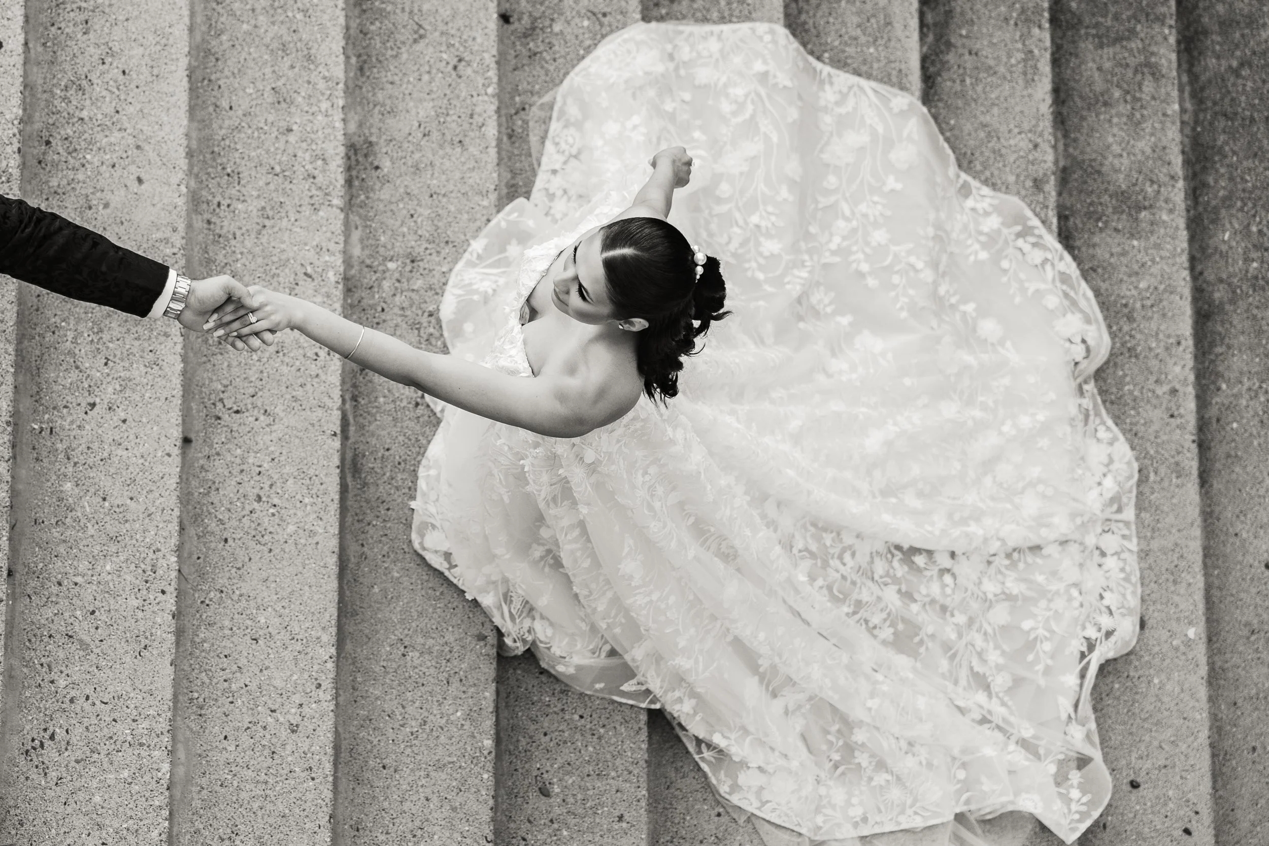 Overhead black and white photo of a bride in a wedding gown holding hands with a groom dressed in a suit, standing on outdoor steps.
