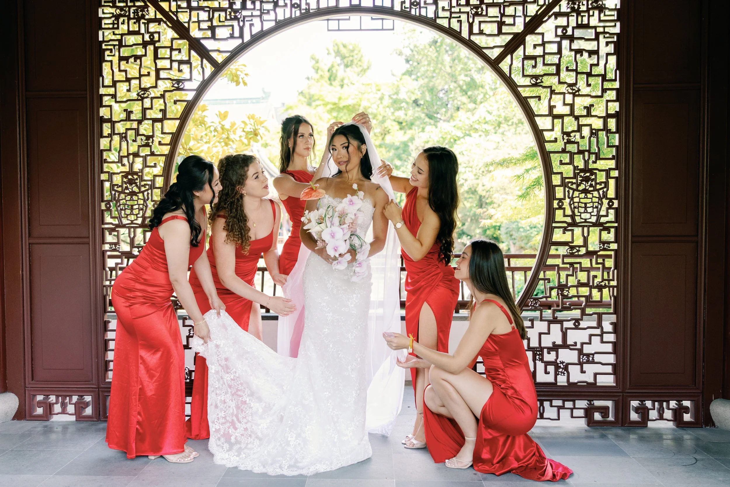 A bride in a white wedding dress holding a bouquet of white and pink flowers, surrounded by seven bridesmaids in red dresses, one kneeling and the others standing, all helping the bride with her veil, in front of a decorative circular window with a g