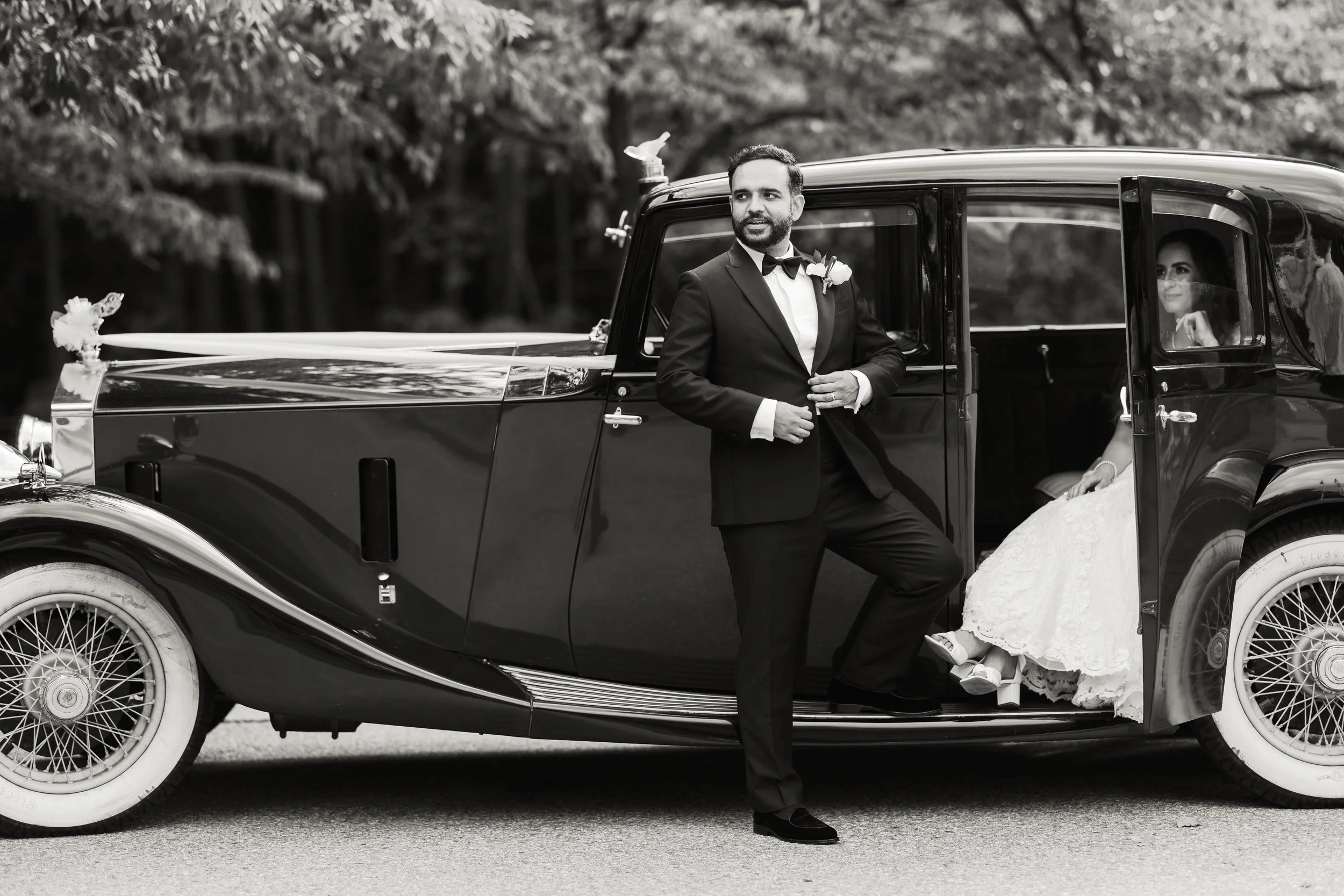 A groom in a tuxedo standing beside a vintage car with a bride sitting inside. The bride is wearing a wedding dress and veil, and the groom is smiling with one foot on the ground. The car is decorated for a wedding, with trees in the background.