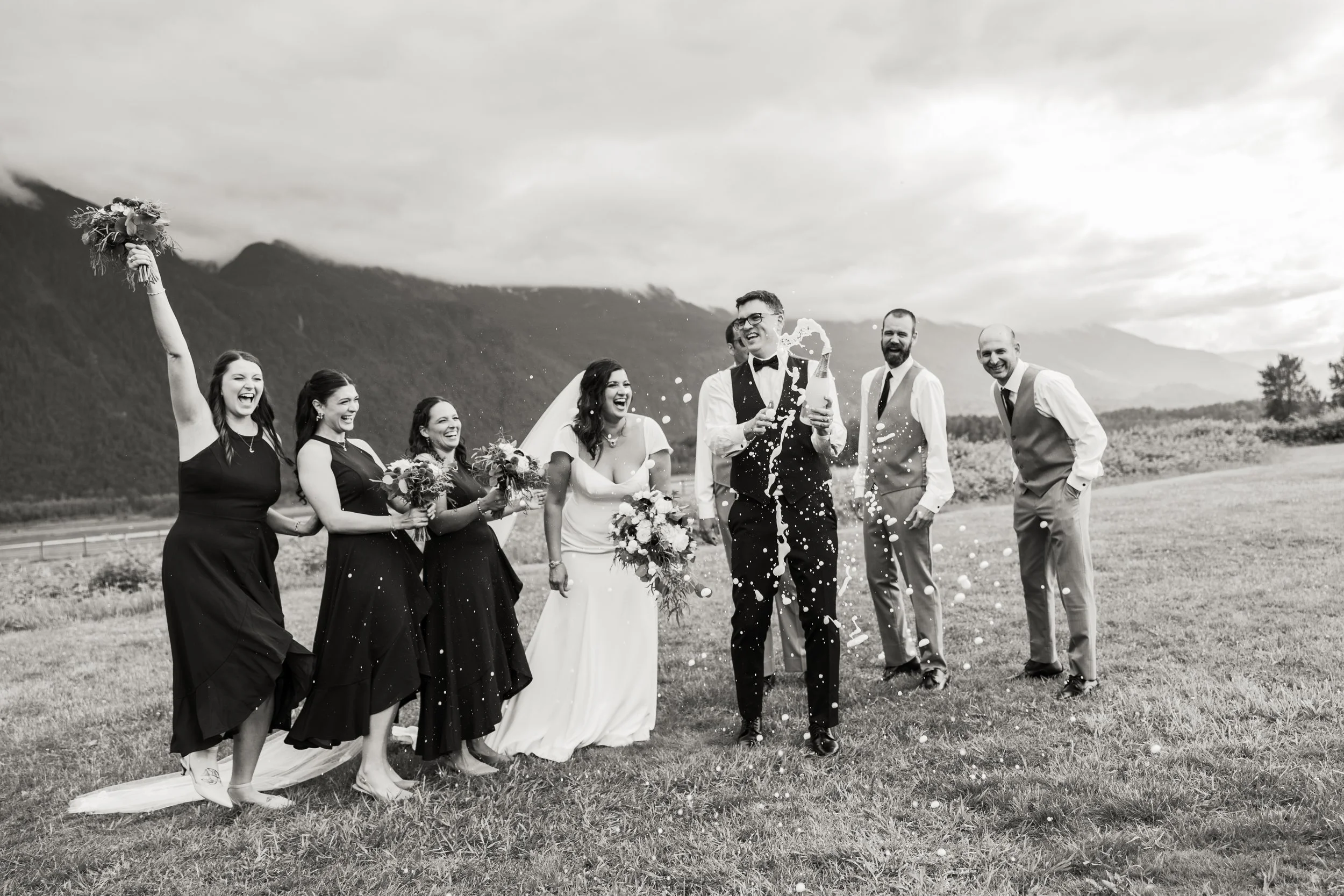 A wedding celebration outdoors with a bride, groom, bridesmaids, and groomsmen, where the groom is opening a champagne bottle, and the group is cheering with mountains in the background, in black and white.