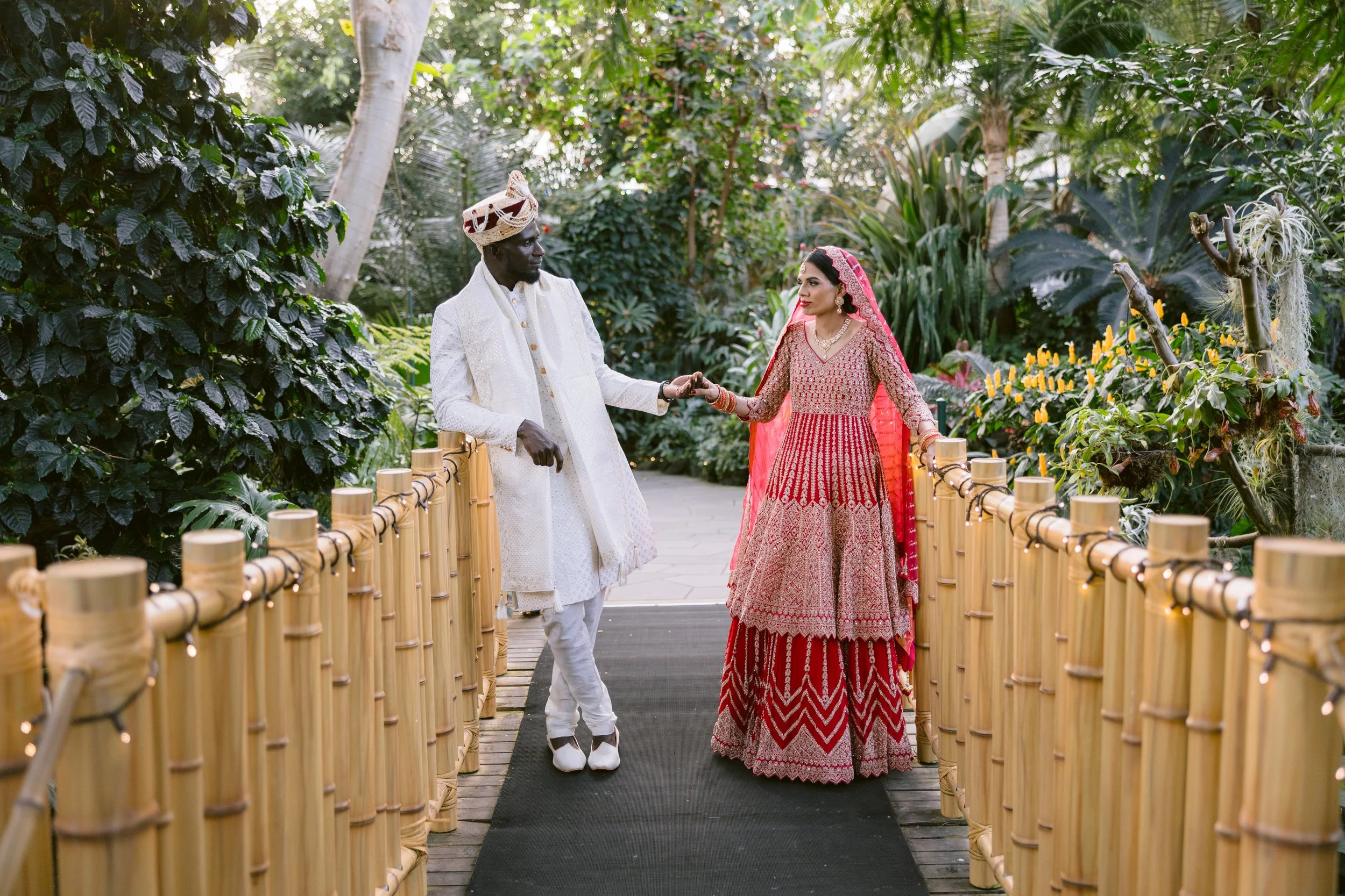 A bride and groom in traditional Indian wedding attire holding hands on a bamboo bridge surrounded by lush green plants in Bloedel Conservatory.